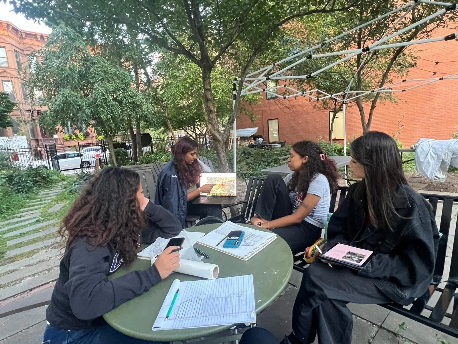Four young women sitting at a round outdoor table, engaged in a discussion in a park with trees, benches, and a brick building in the background.