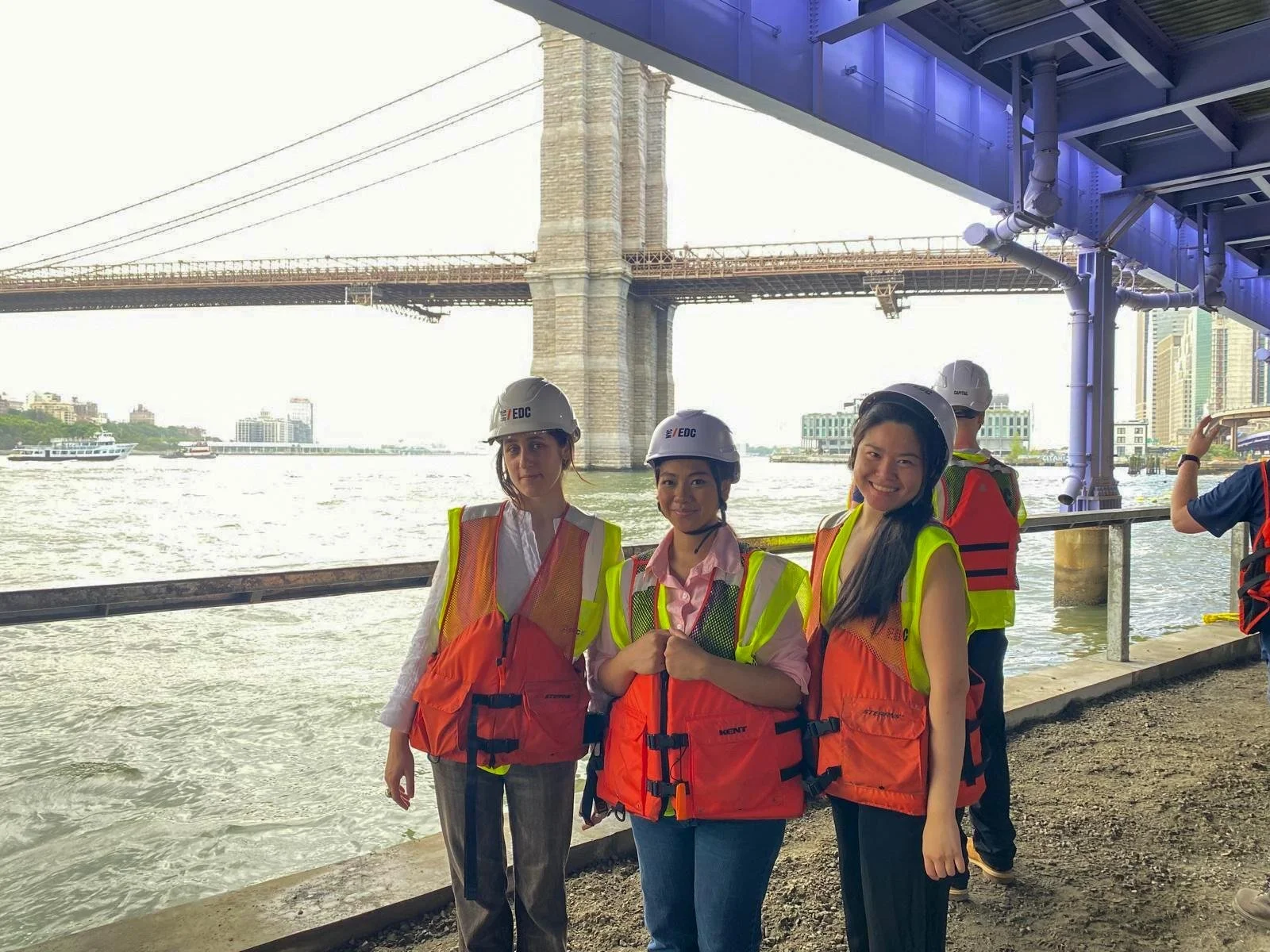 Three women wearing safety helmets and orange life jackets standing under a bridge near a river, with a city skyline in the background.