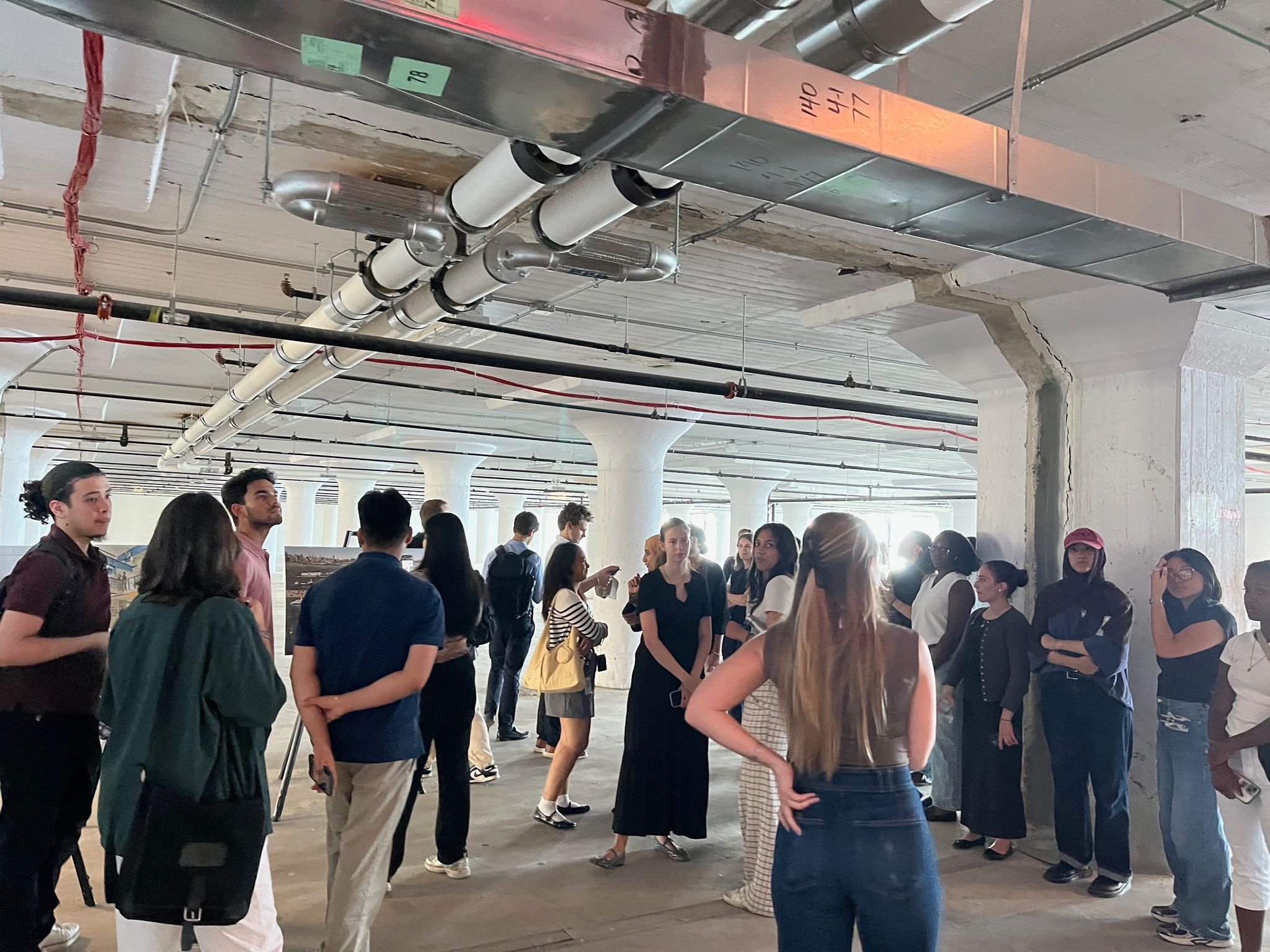 A group of people gathered in an industrial-style space with exposed concrete pillars and ceiling pipes, listening and talking to each other.