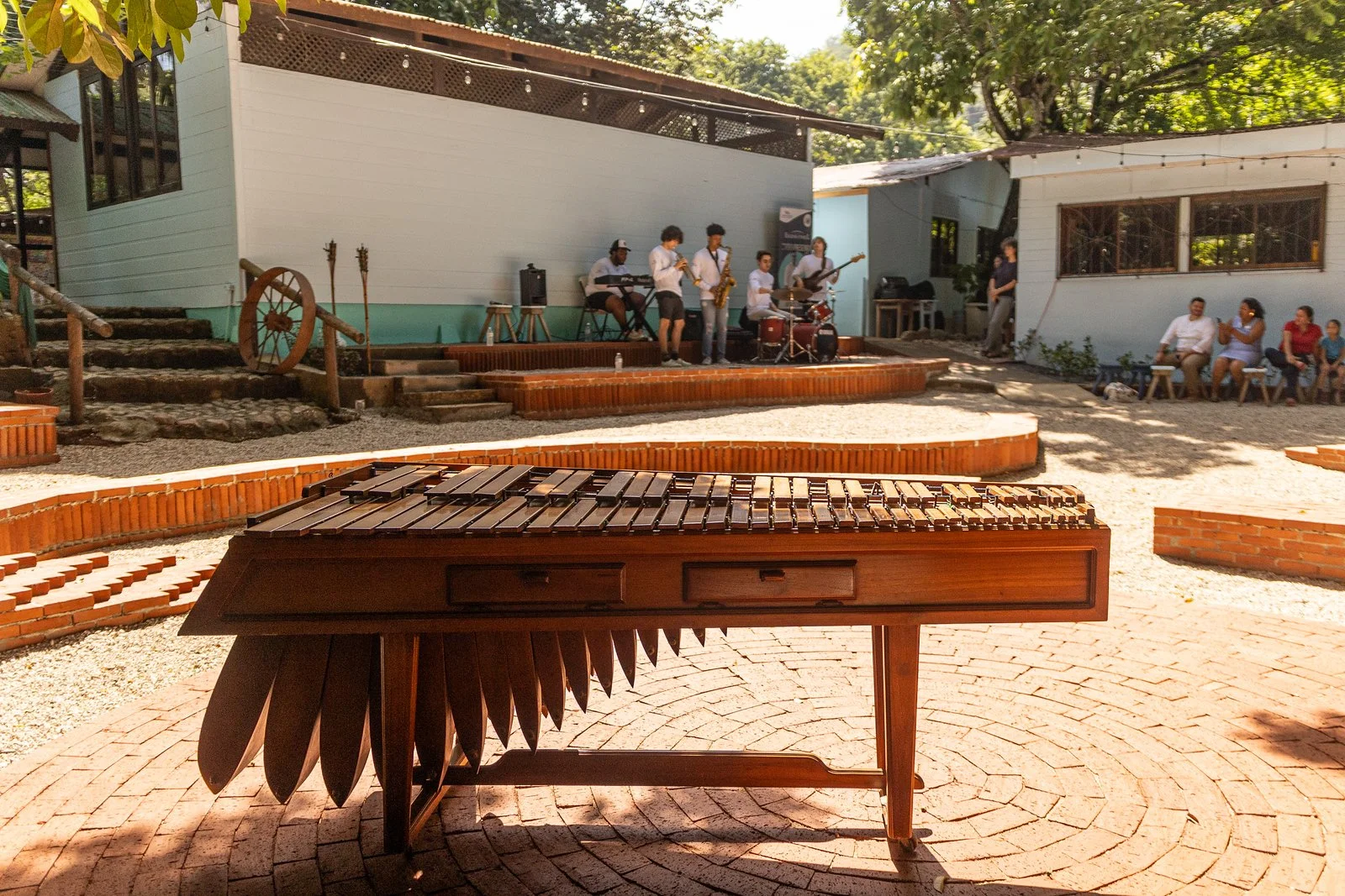 An outdoor stage with a band performing, and a wooden marimba in the foreground. Audience members are seated on benches to the right, and there are trees and buildings surrounding the scene.