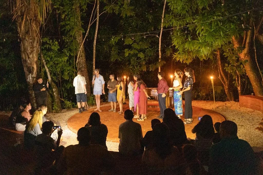 A group of people is on a small outdoor stage surrounded by trees, performing with candles and a small audience watching during nighttime.
