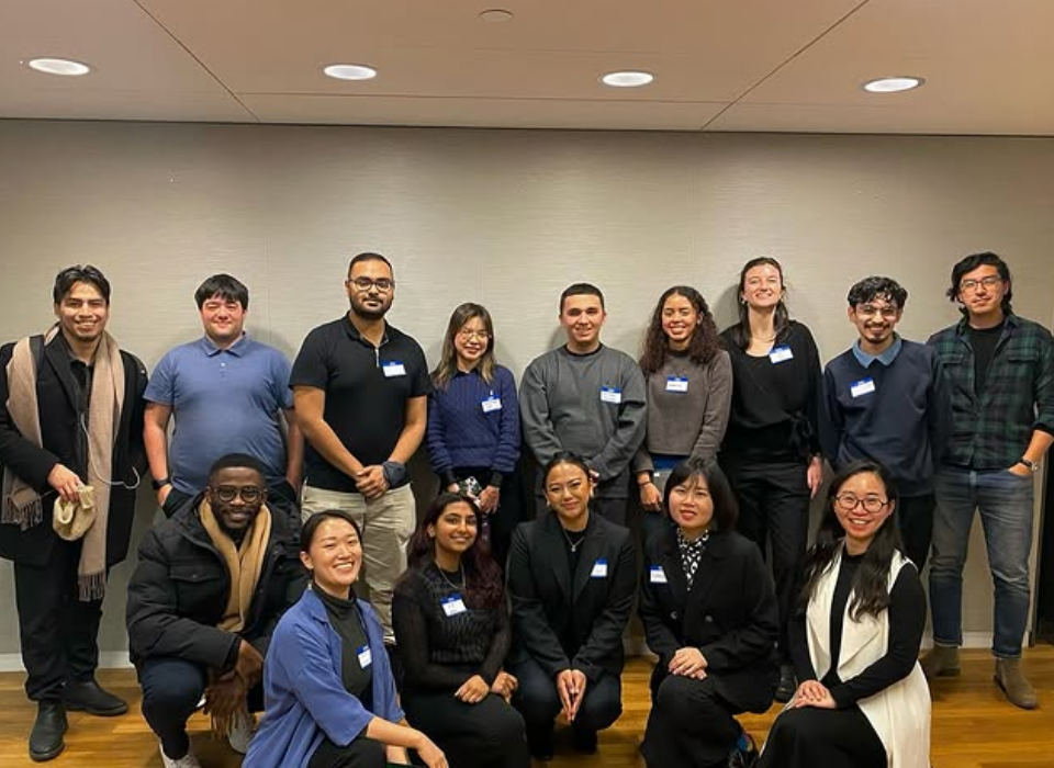 Group of fifteen diverse young adults posing indoors against a neutral wall, some standing and some kneeling, smiling for the camera at a social or professional event.