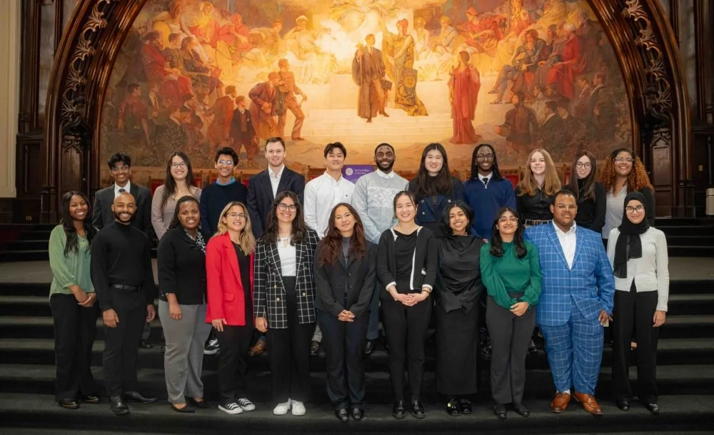 A diverse group of young adults and professionals standing on a stage with a large, ornate mural in the background, posing for a group photo in a formal setting.