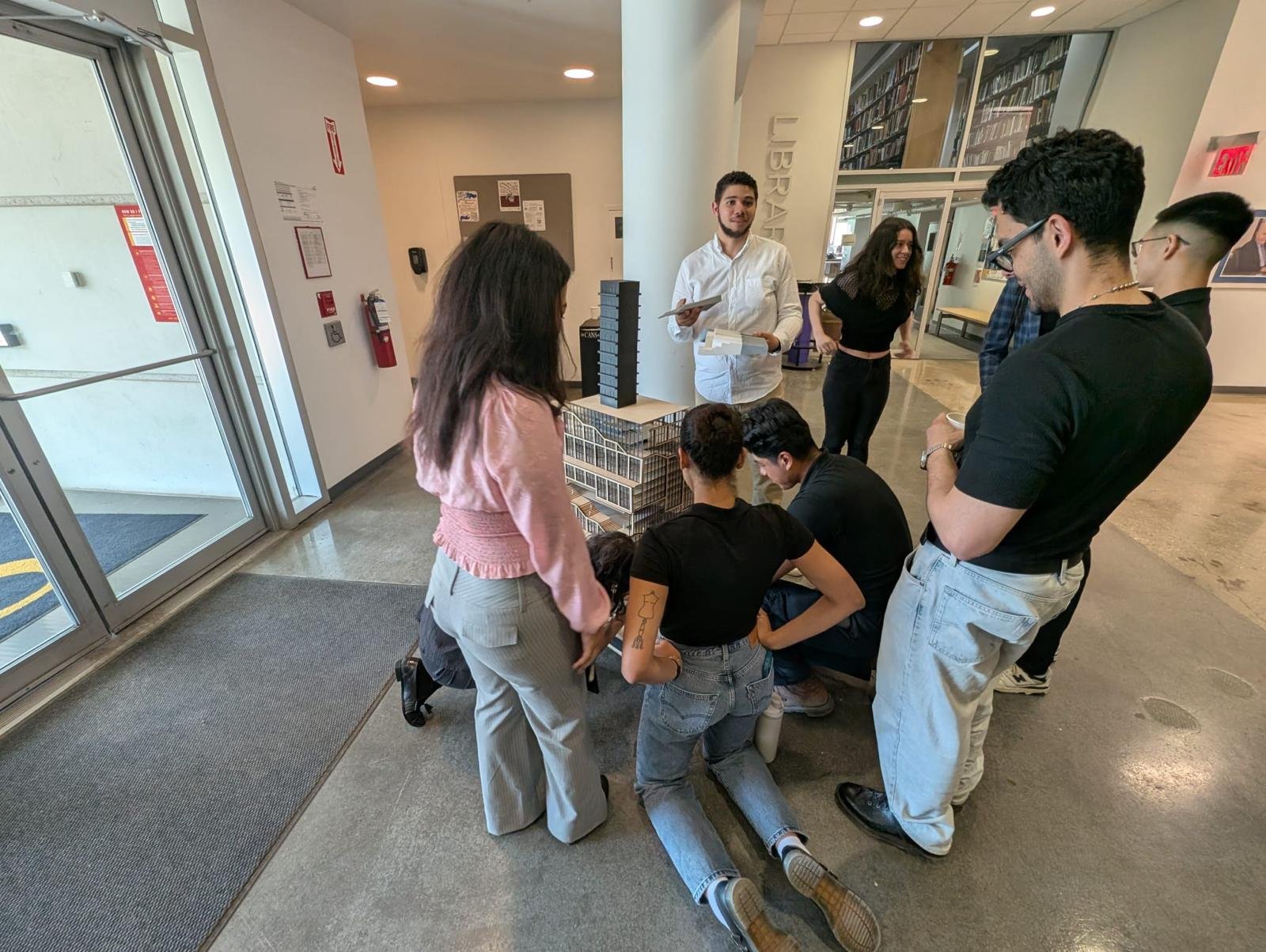 A group of people gathered around a scale model of a building inside a library, engaging in a discussion.