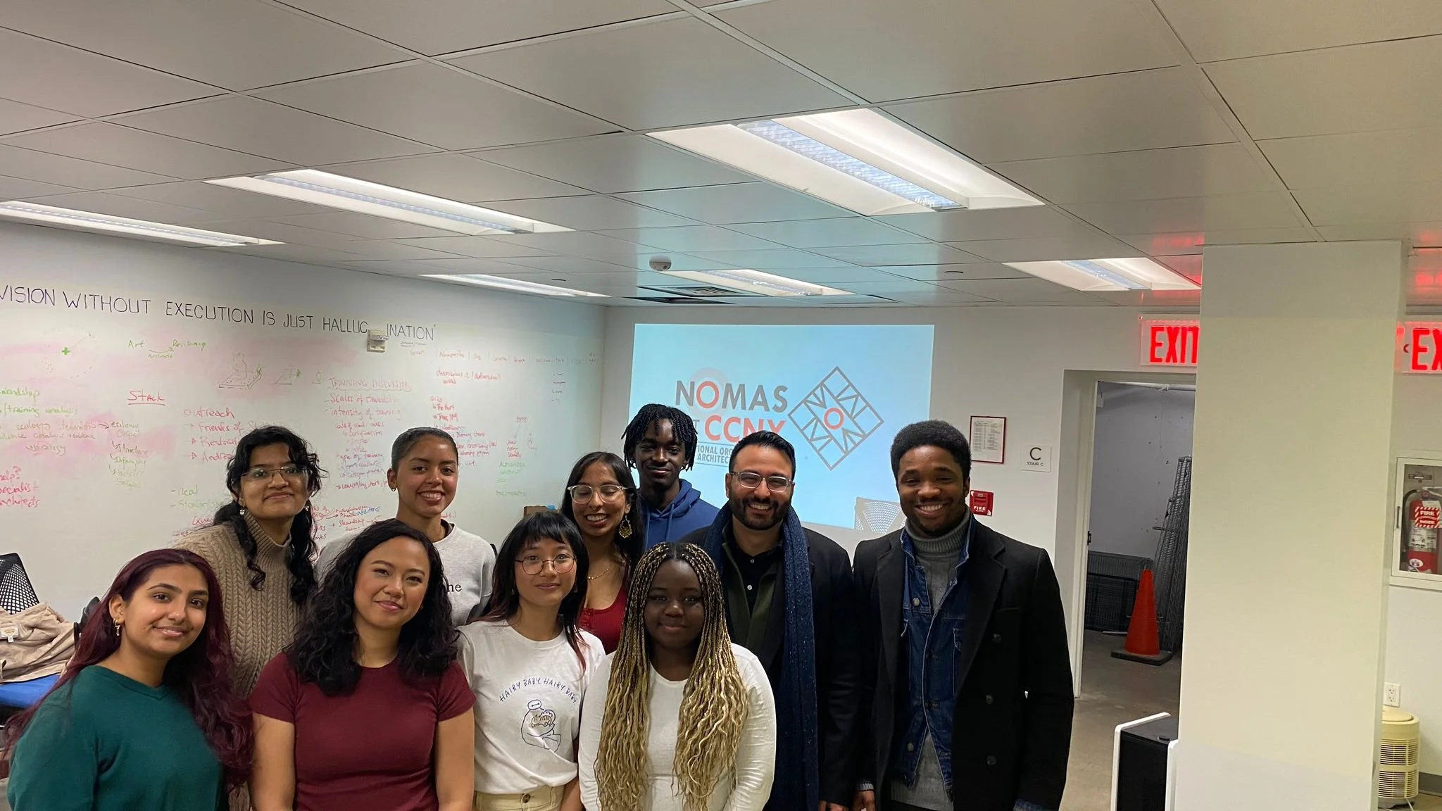 A group of ten diverse individuals standing together in an office or classroom, smiling at the camera. In the background, there is a whiteboard with handwritten notes and a projector screen displaying the logo for NOMAS CCNY, part of the National Org