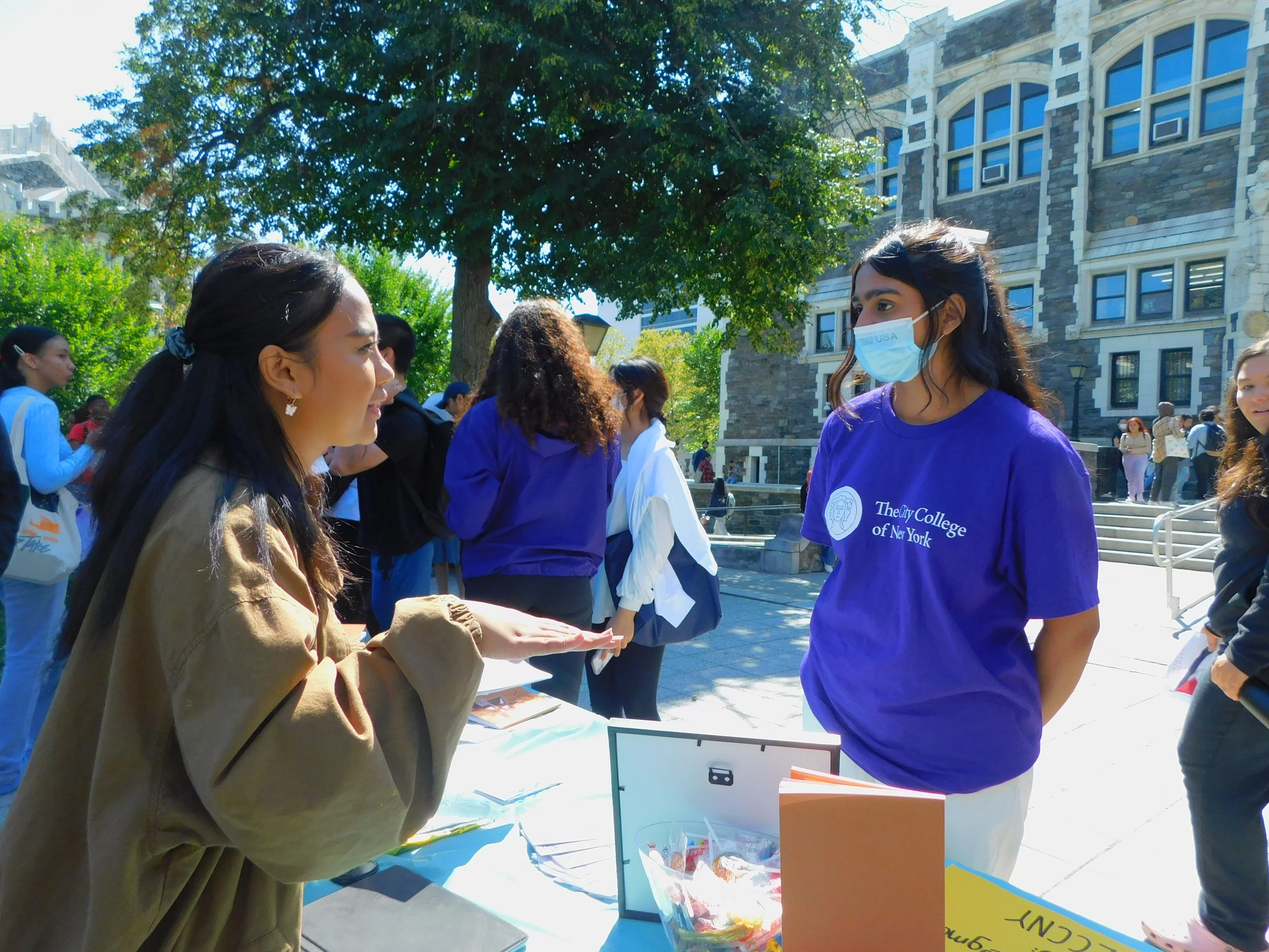 Two young women having a conversation outdoors at a college event. One woman is wearing a beige jacket and gesturing with her hands, the other woman is wearing a purple t-shirt with the college's name and a face mask.