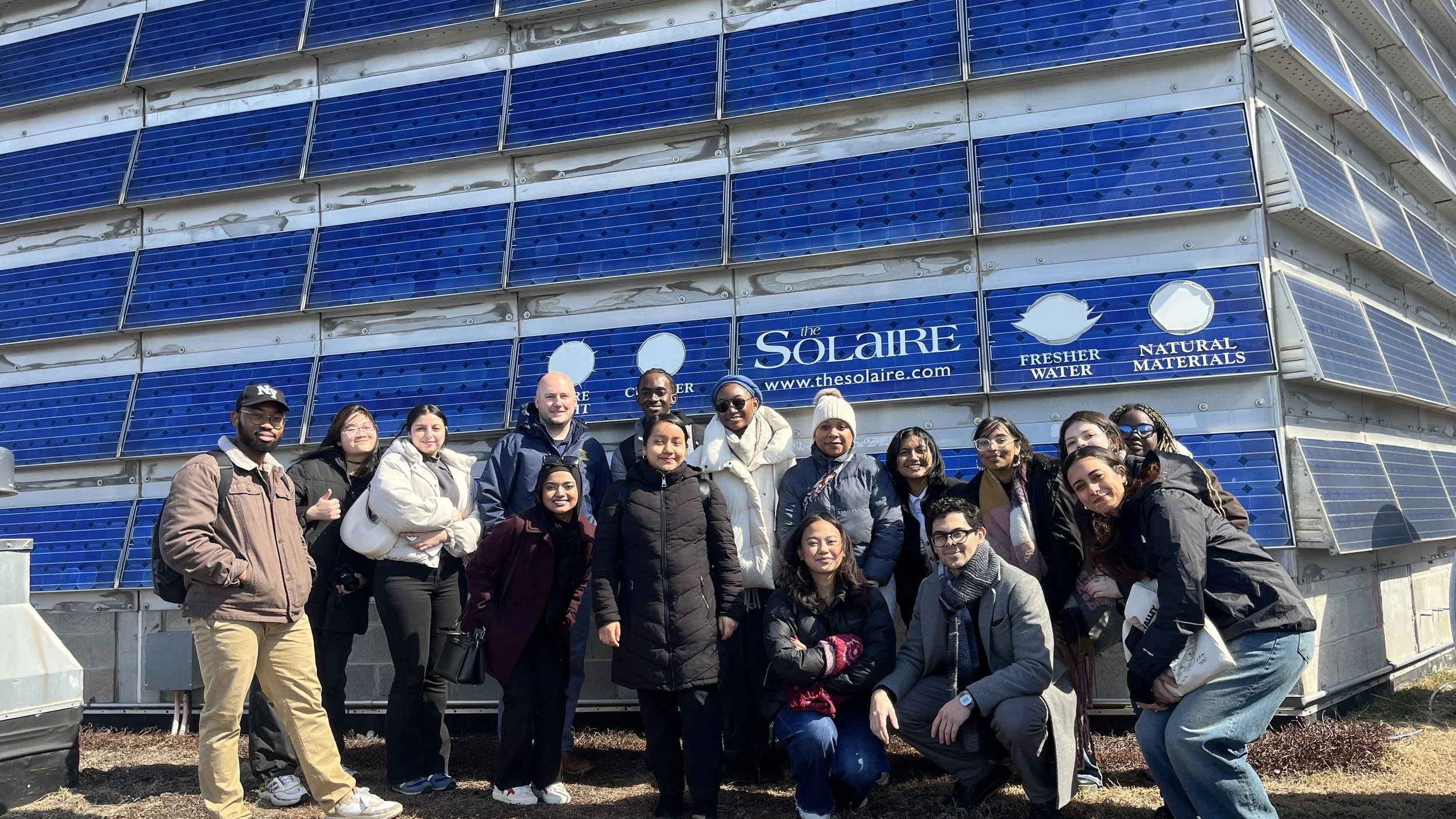 Group of people posing outdoors in front of a building covered with blue solar panels, some wearing jackets and sunglasses.