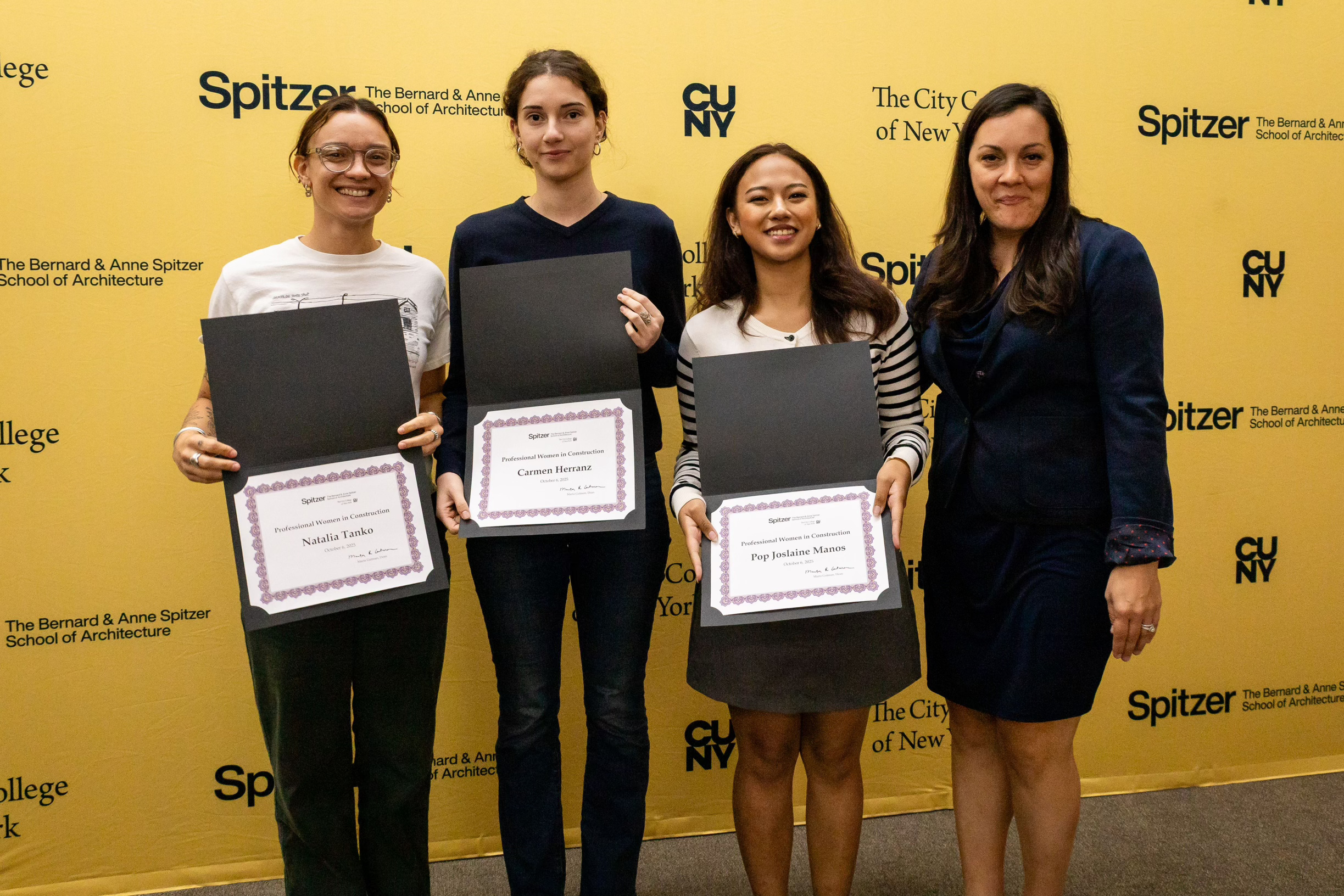 Four women standing in front of a yellow backdrop with black text and logos, holding certificates, smiling for the photo.