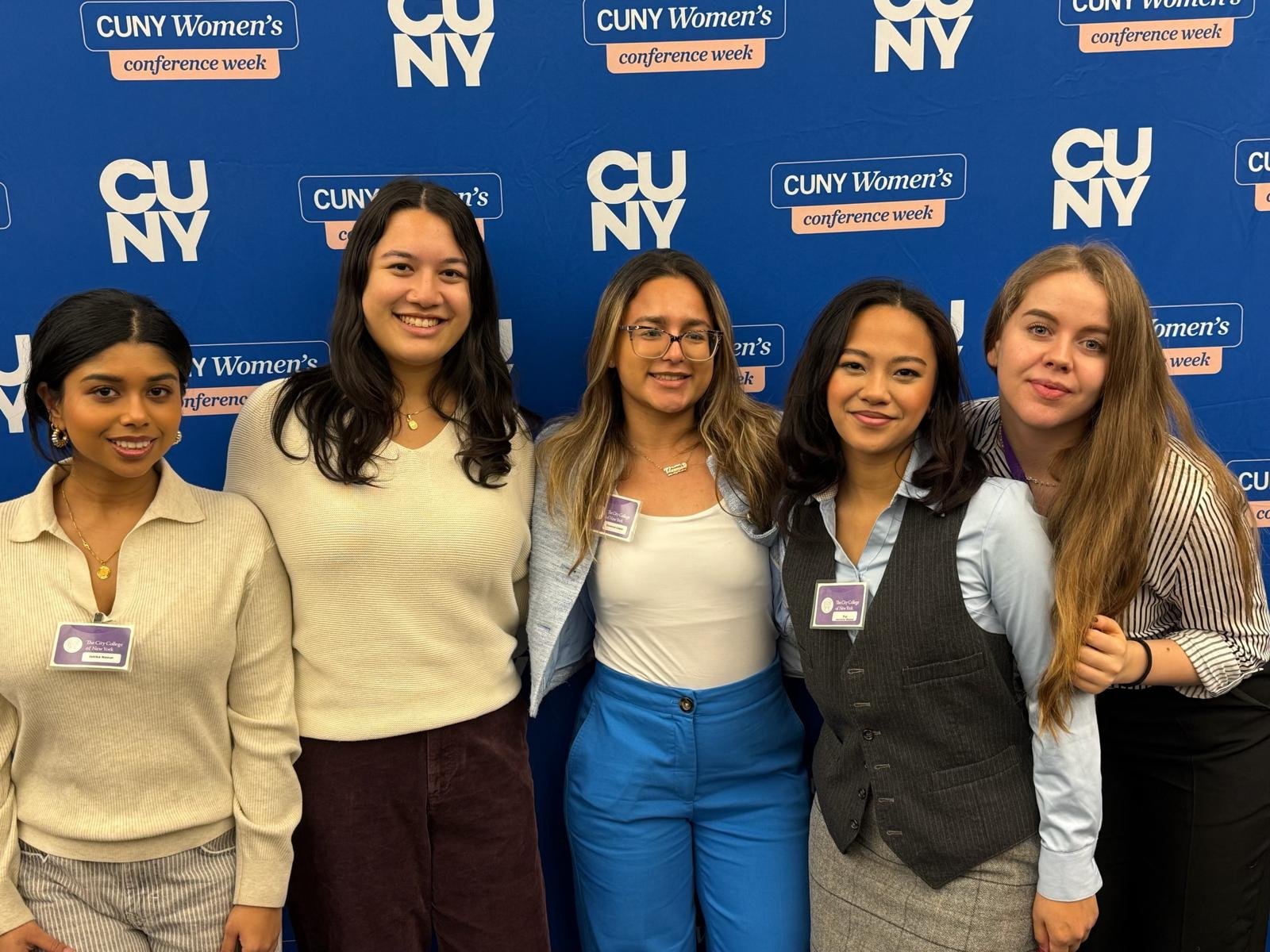 Five women standing together in front of a blue backdrop with CUNY Women's Conference Week logo, smiling at the camera.