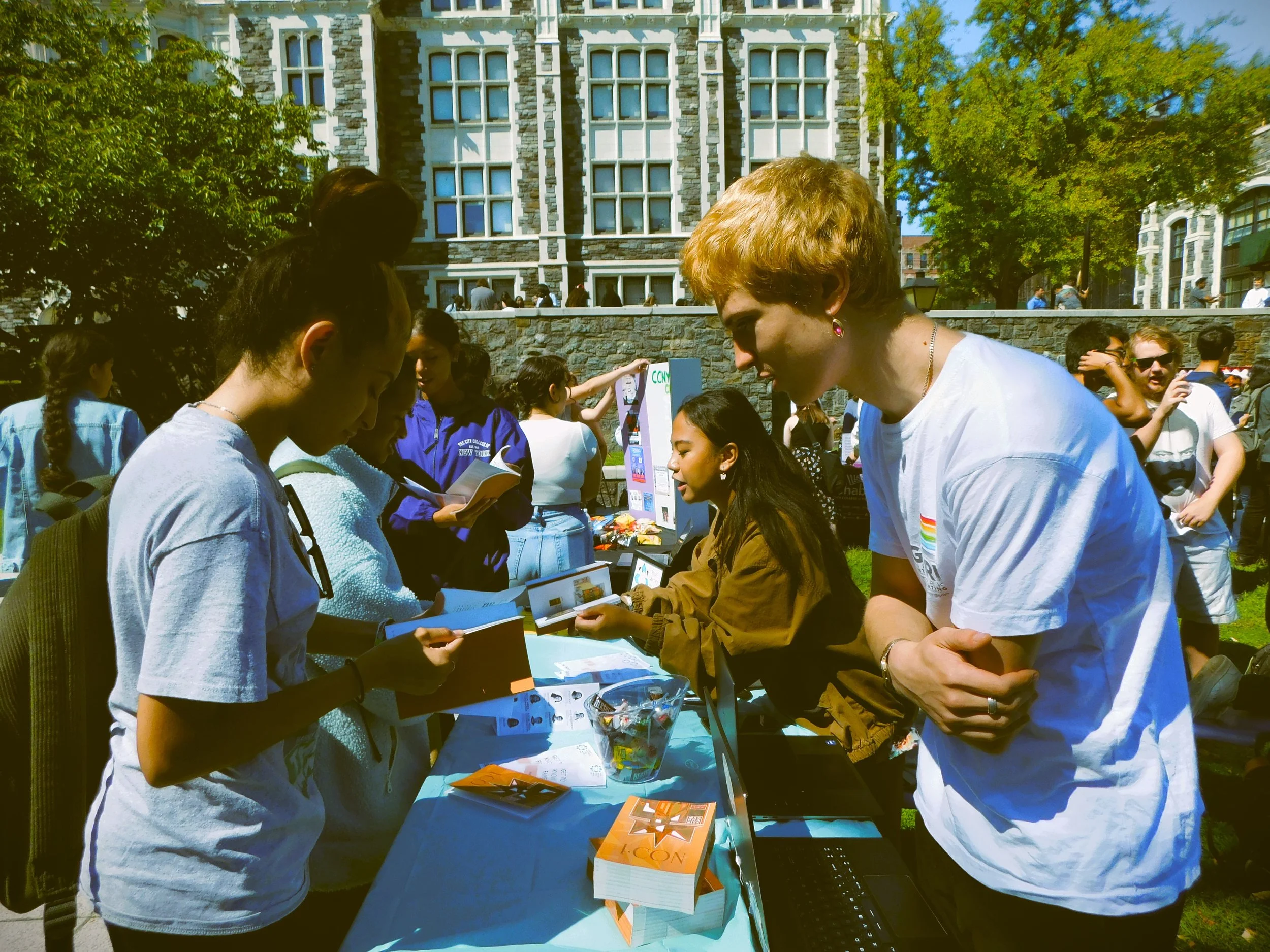 People gathered around an outdoor table at a public event on a sunny day, with a historic building in the background.