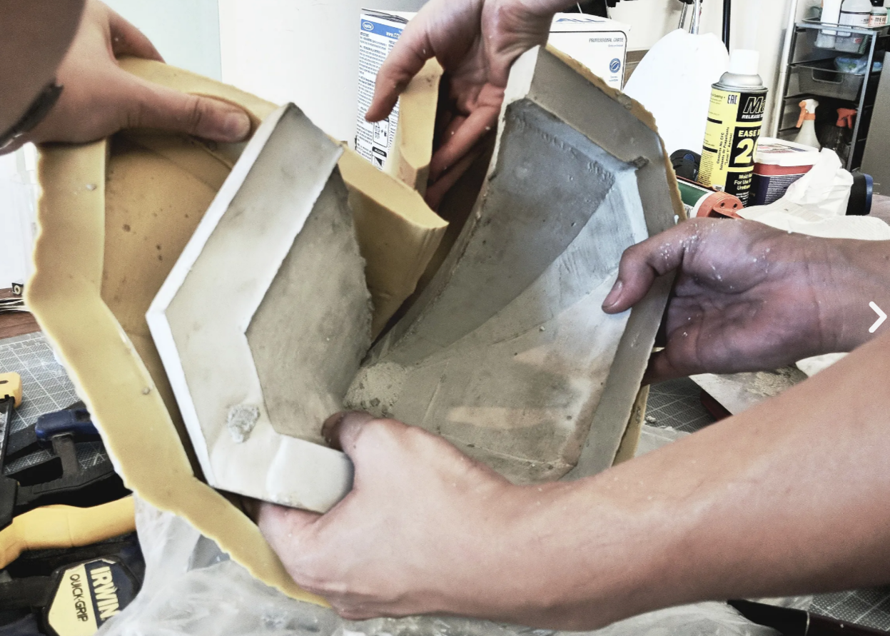 Two people are handling a mold or negative in a workshop, with tools, adhesive, and supplies visible in the background.