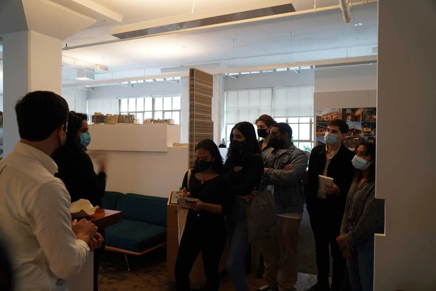 A group of people wearing face masks inside an office or gallery space, listening to a person at the front, some taking notes and holding bags.