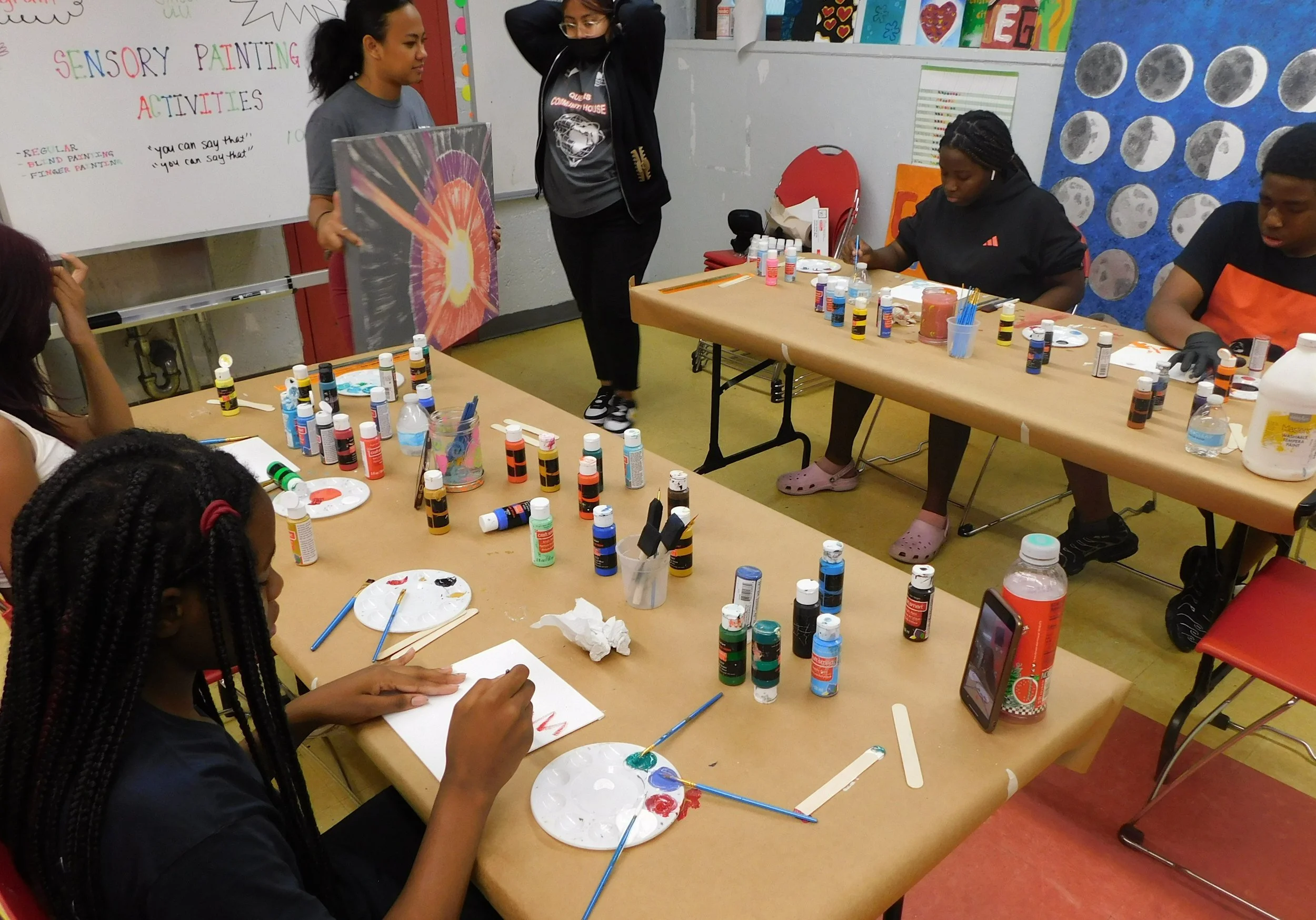 A group of children and two adults participating in a sensory painting activity in a classroom. The table is covered with paint bottles, brushes, and palettes, with some kids painting and others observing.