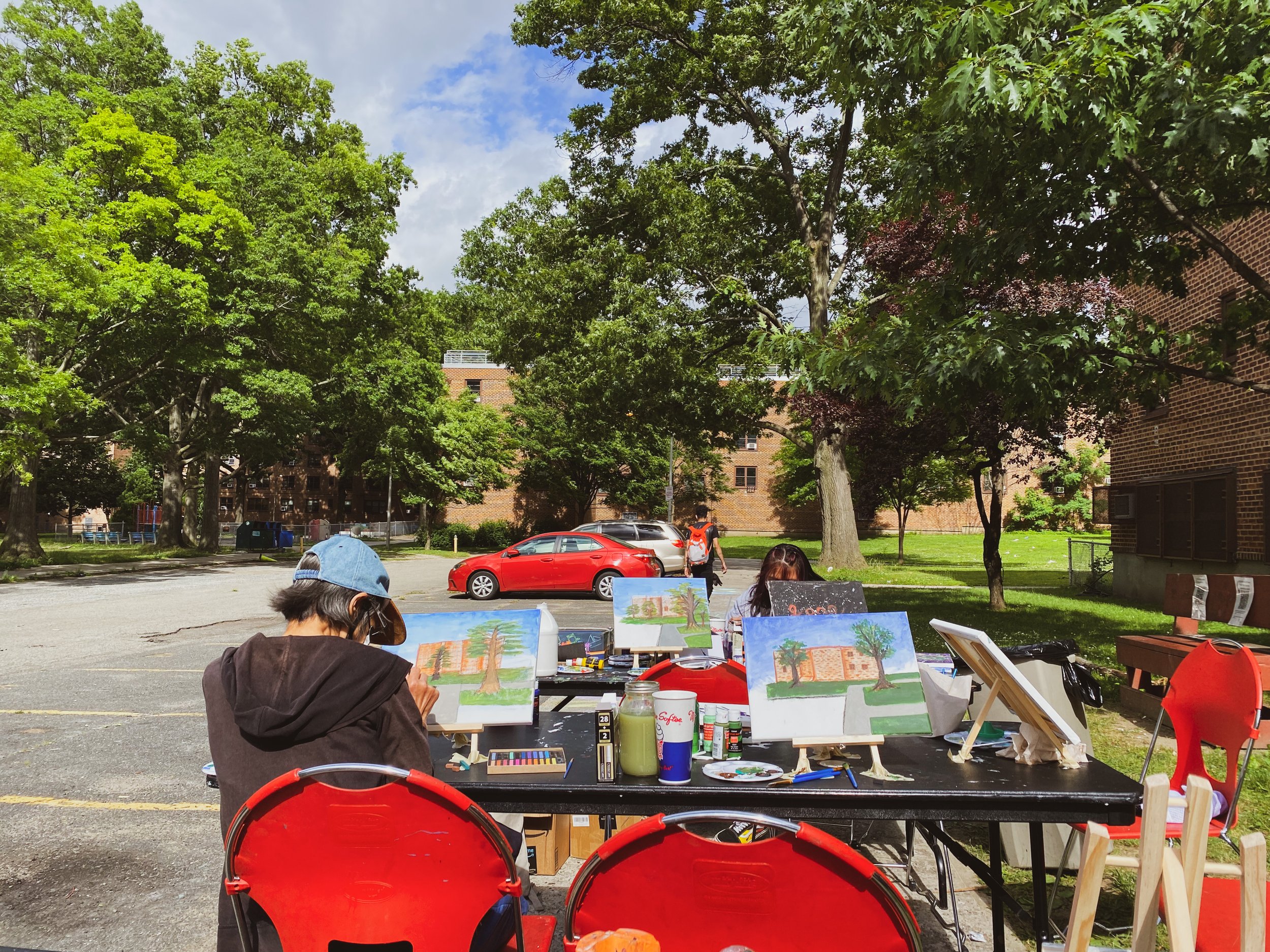 People painting on outdoor easels in a parking lot surrounded by trees and brick buildings, with a red car parked nearby.