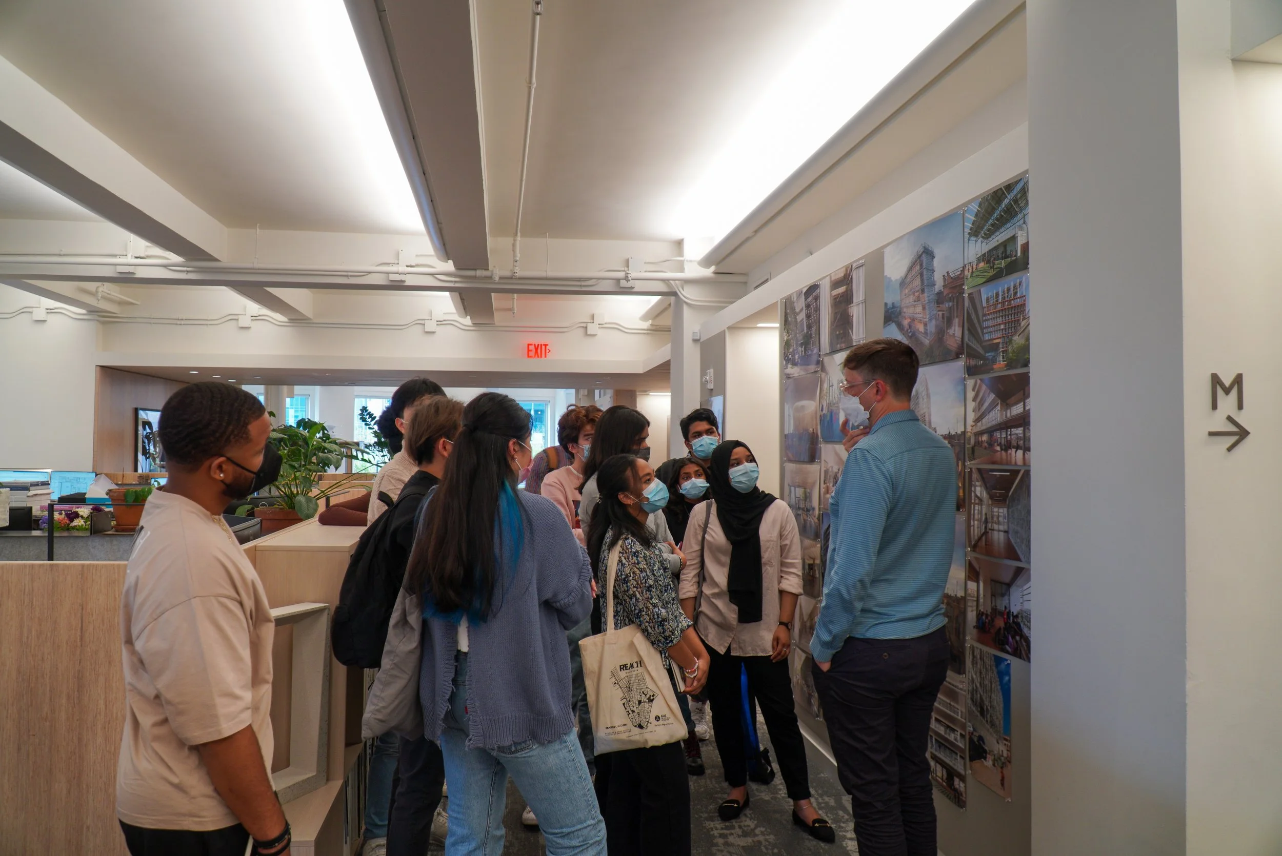 A group of people wearing masks listening to a man in a blue shirt who is explaining architectural images on a wall.