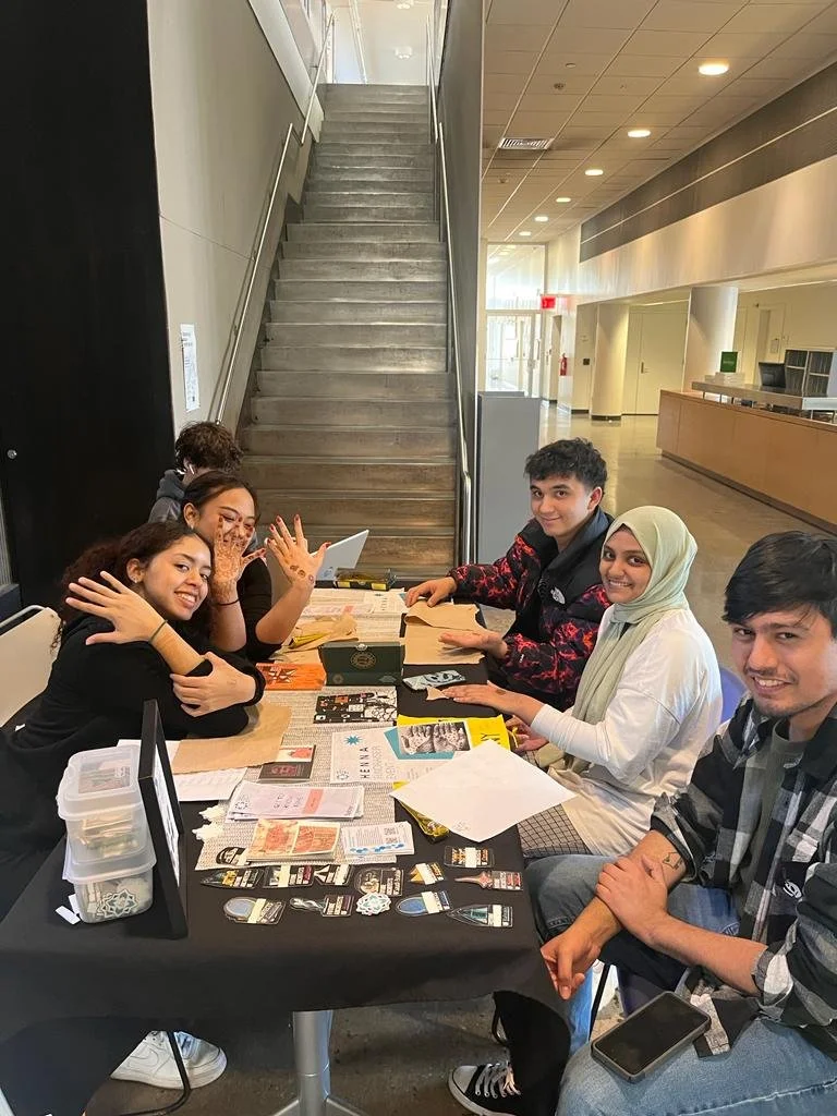 A group of five young adults sitting around a table in a well-lit indoor space, engaging with various printed materials, with a staircase and hallway in the background.