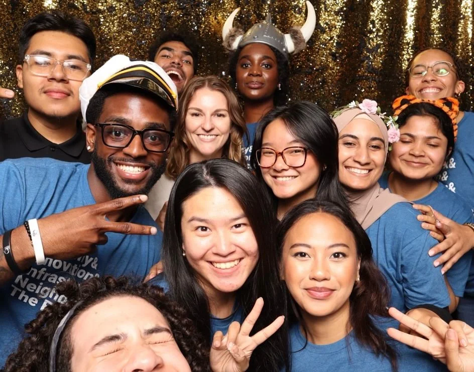Group of diverse friends smiling and posing together at a celebration, some wearing costumes or accessories like a Viking helmet, flower crown, and tiger headband, in front of a gold glittery backdrop.