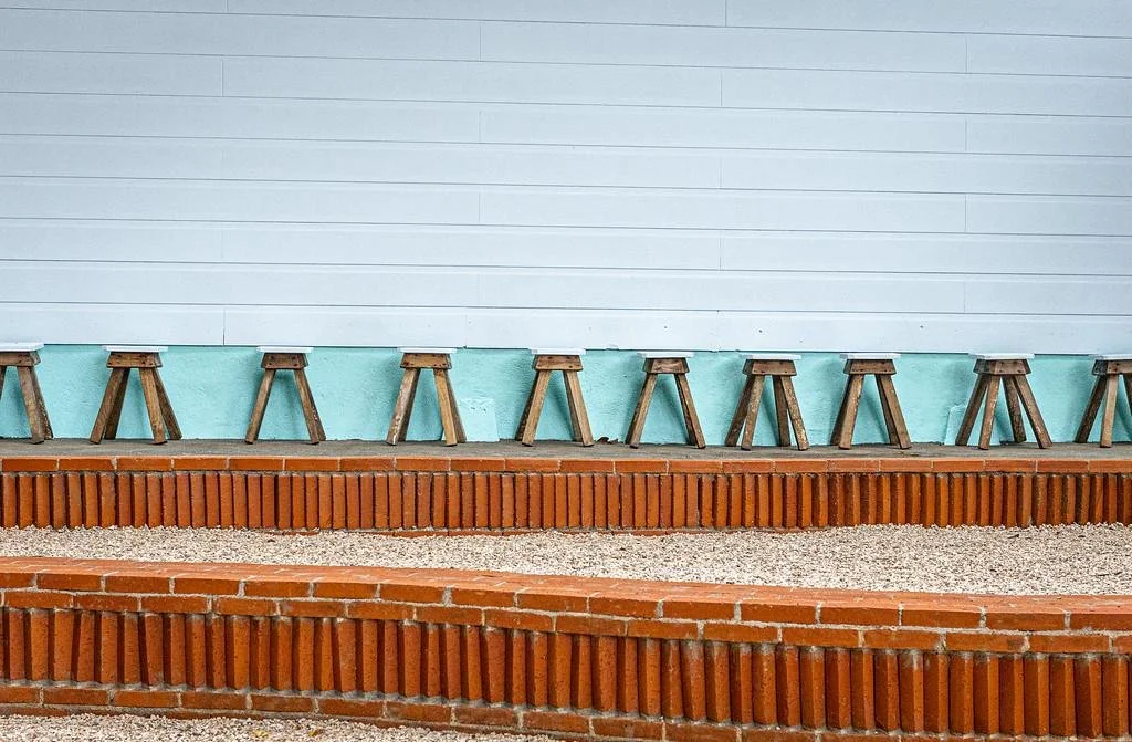 Row of small wood stools lined up against a white wall with green trim and brick foundation, outdoors.