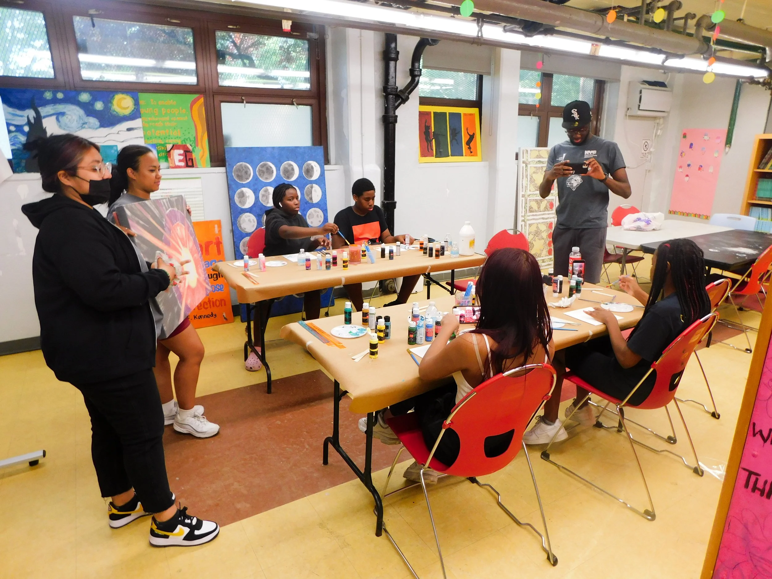 A group of children and teens participating in an arts and crafts activity in a colorful classroom. Some are seated at tables with paint bottles and brushes, while others stand or watch. A man is standing, looking at his phone. There are colorful pos