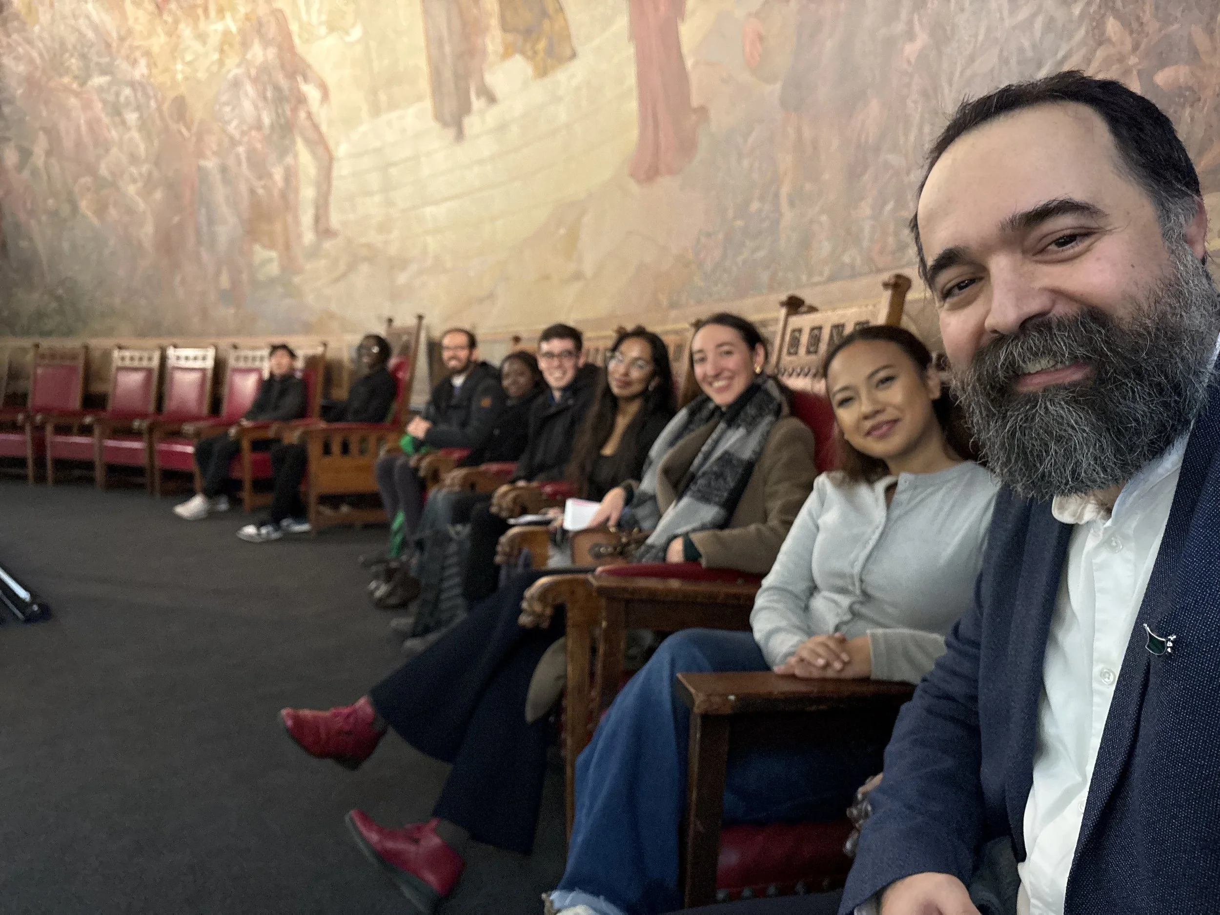 A group of diverse people sitting in a row on wooden chairs with red upholstery in a large room with a mural on the wall, taking a selfie.