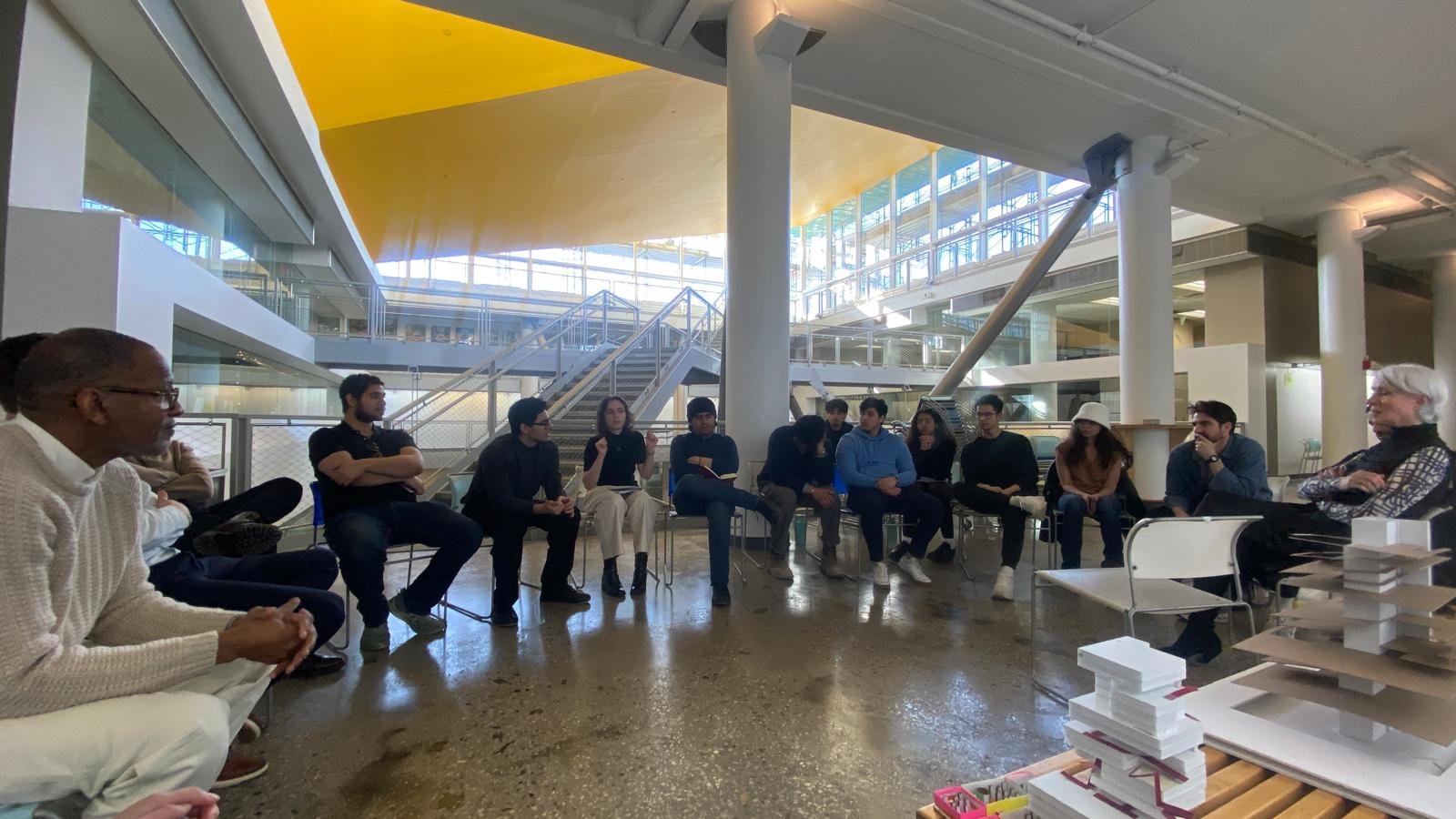 A group of people sitting in a circle in an indoor space with modern architecture, skylights, and a yellow ceiling, engaged in a discussion or meeting.