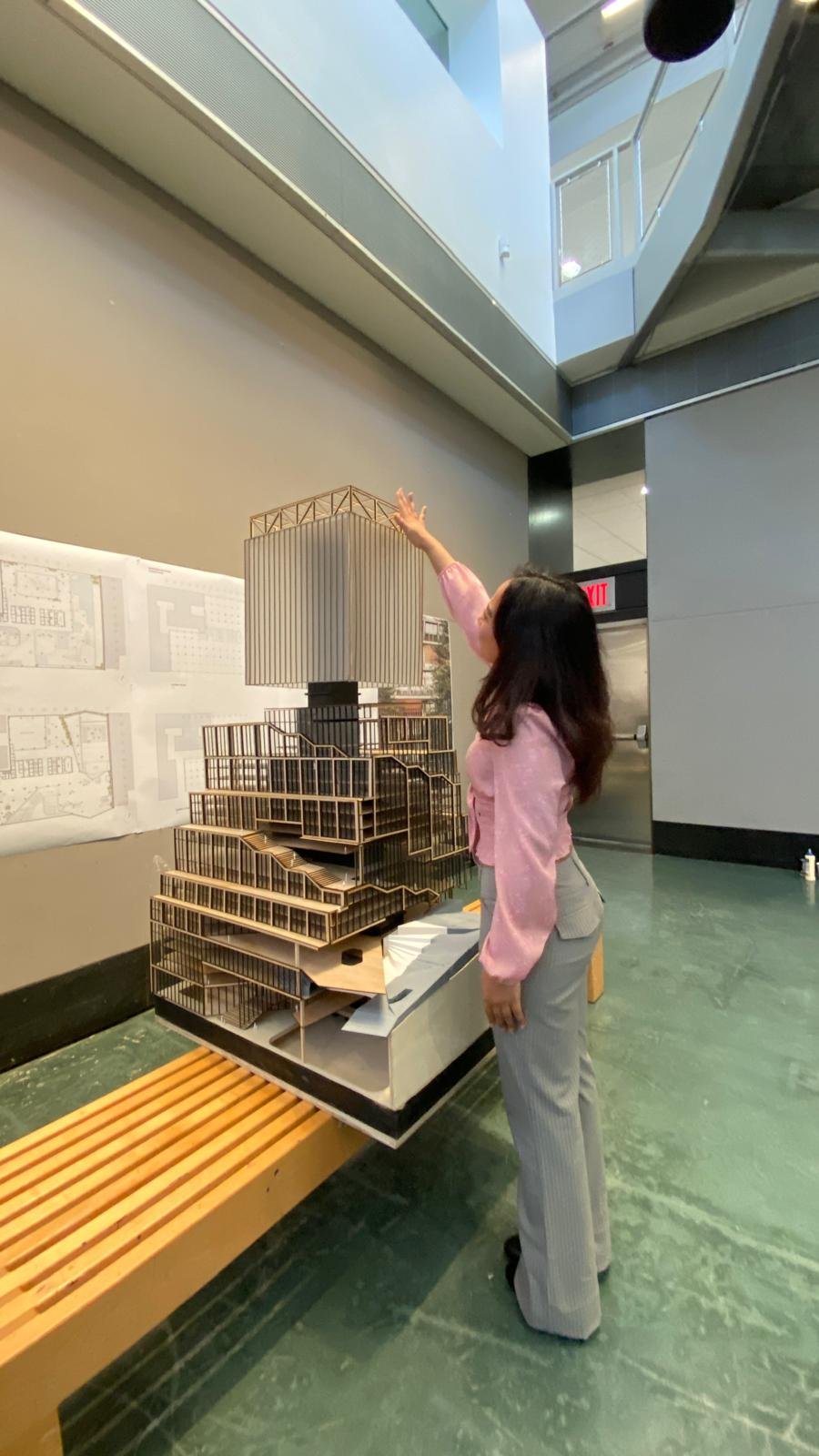 A woman with long dark hair in a pink jacket and striped pants is pointing at a scale model of a multi-story building with a unique stepped design, displayed on a table. She is indoors, near a beige wall with architectural plans, in a modern building
