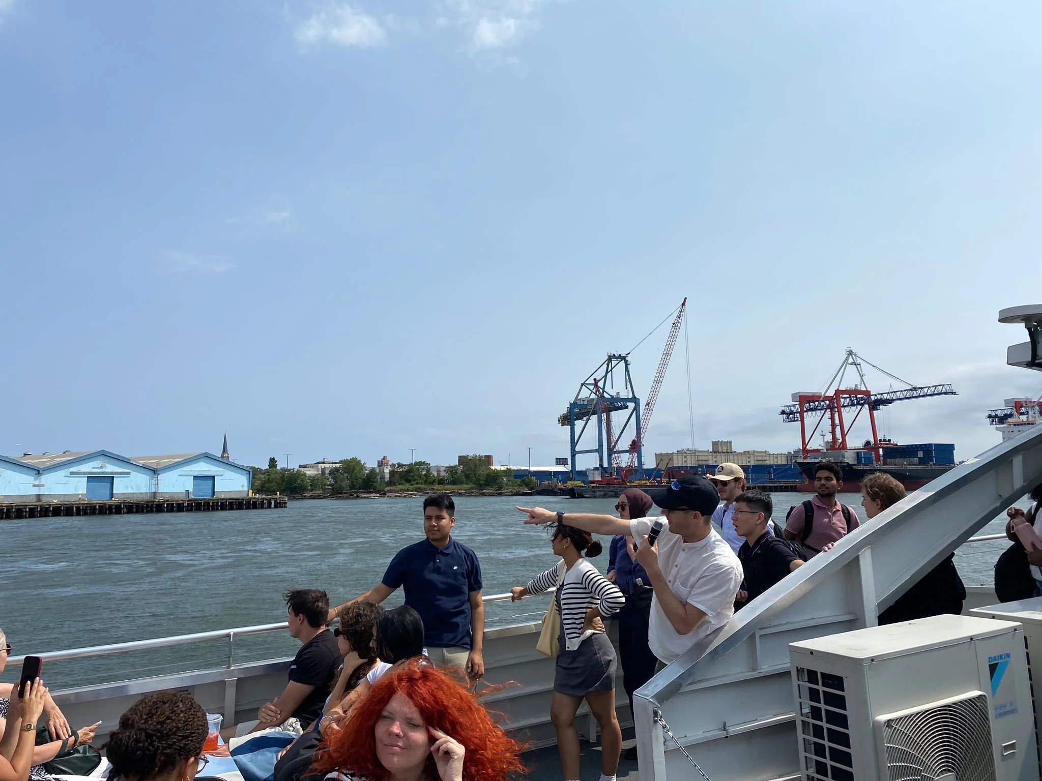 A group of tourists on a boat tour, with some taking photos and others listening to a guide pointing towards a port with cranes and industrial buildings in the background during daytime.