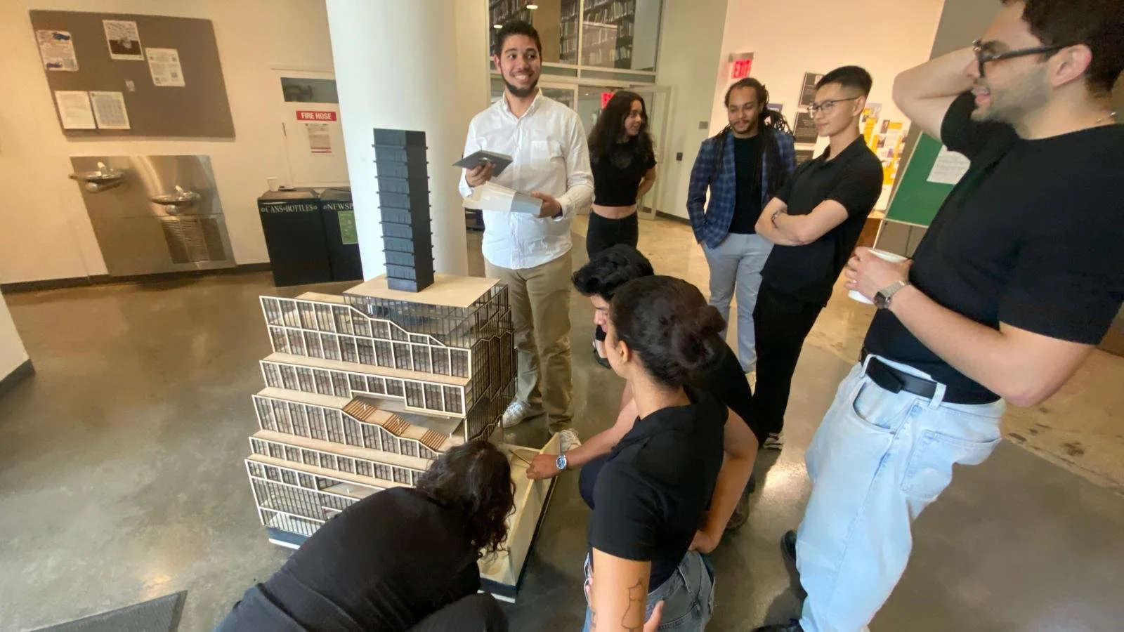 Group of seven people gathered around a detailed architectural model of a building in an indoor setting, with a man in a white shirt holding documents, engaging with the group.