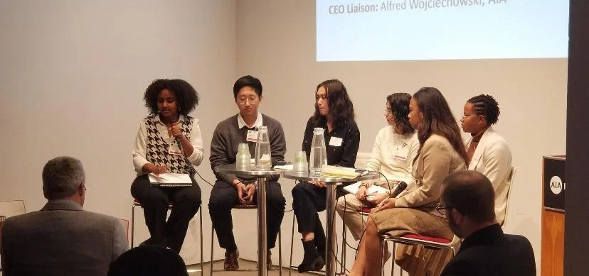 Six diverse women seated on a panel during a discussion, with two men and a woman in the audience listening.