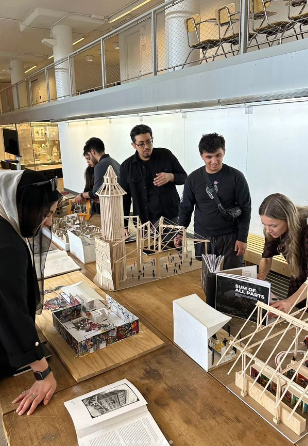 Group of young adults gathered around a table with architectural models and books, working on a project in a well-lit indoor space.