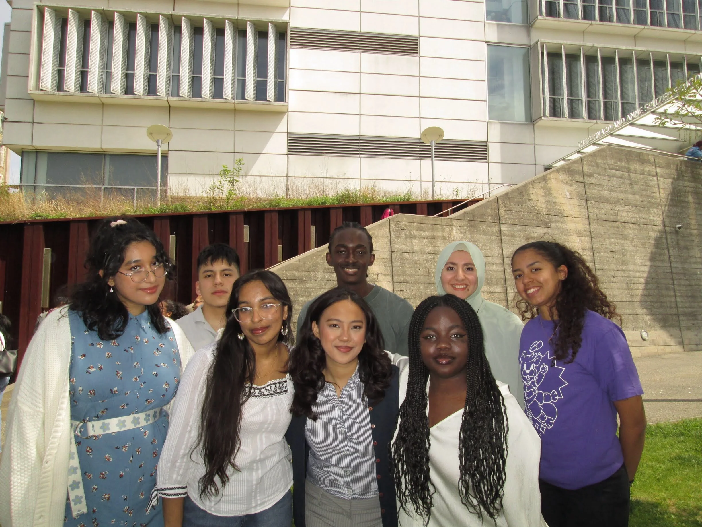 Group of diverse young women and men smiling outdoors in front of a modern building with stairs and greenery.