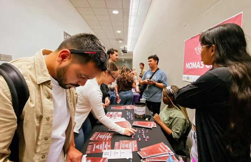 People attending an informational or registration booth at an event, viewing flyers and posters on the table, with a pink sign in the background.
