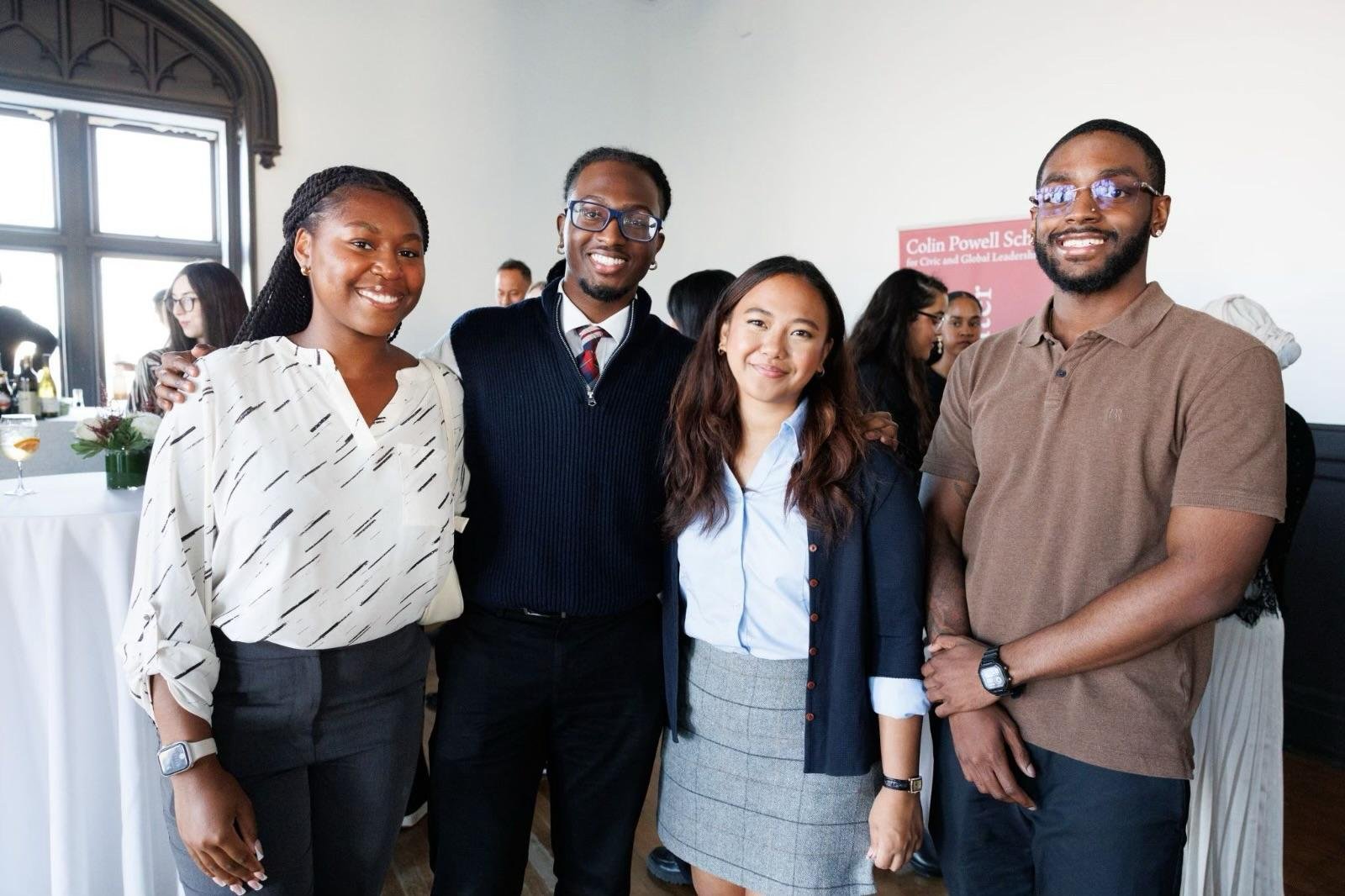 Four diverse young adults standing together indoors, smiling, at a social or professional gathering, with a background of other people and a sign reading "Colin Powell School".