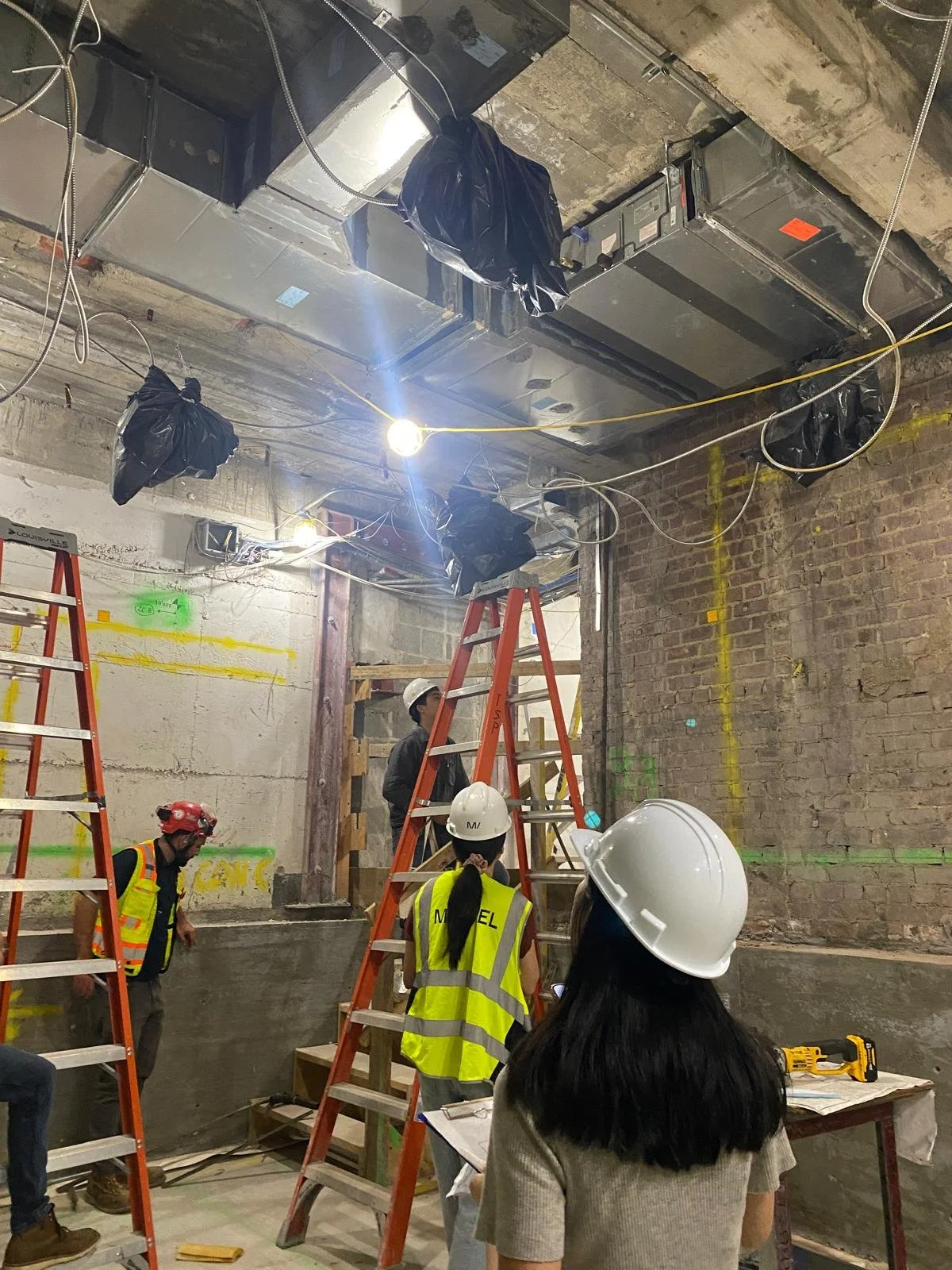 Construction workers with helmets and safety vests working on ceiling installation inside a building with exposed brick walls and ladders.
