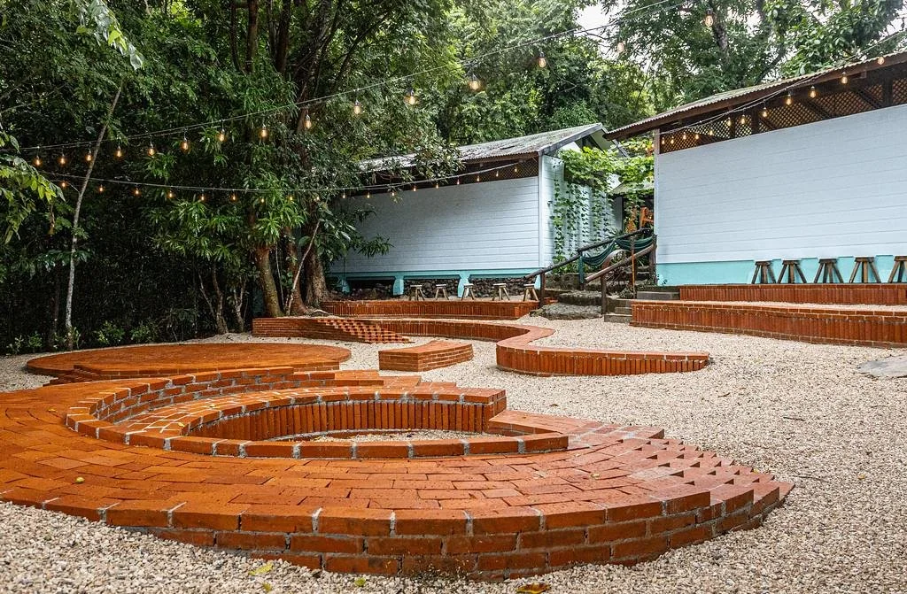 Outdoor patio with circular brick structures on gravel ground, string lights overhead, surrounded by trees and light-blue and white buildings.