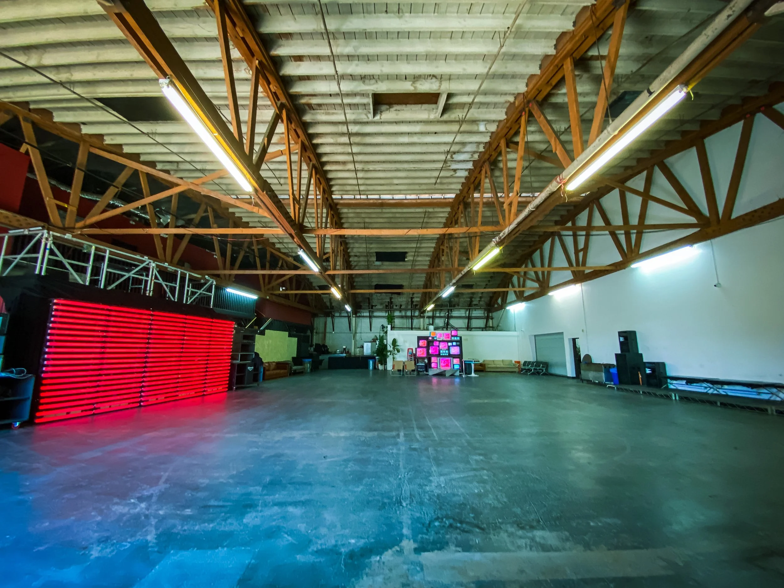 Interior of a large industrial warehouse space with a high ceiling and exposed beams. The floor is concrete, and there's a wall of illuminated red lights on the left side. In the background, a stack of colorful televisions can be seen.