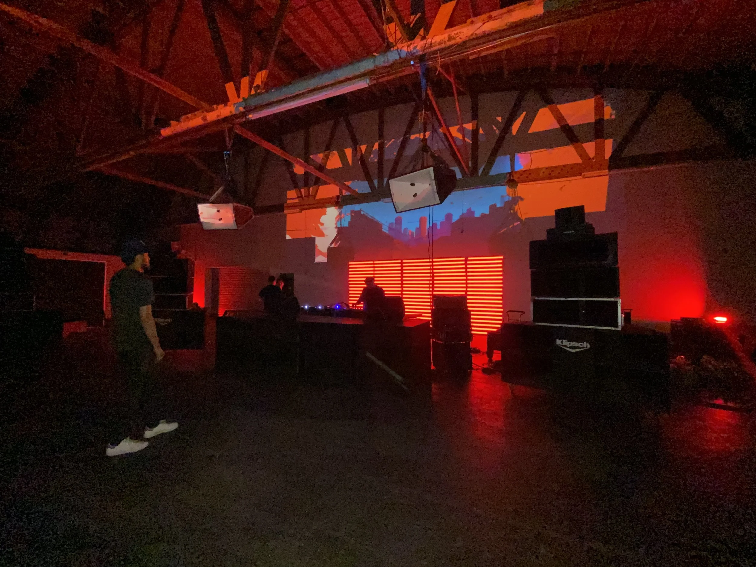 Interior of a dimly lit event space with red lighting, featuring a large sound system, DJ booth, and a projector displaying visuals on the wall. A person stands on the left side observing the setup.