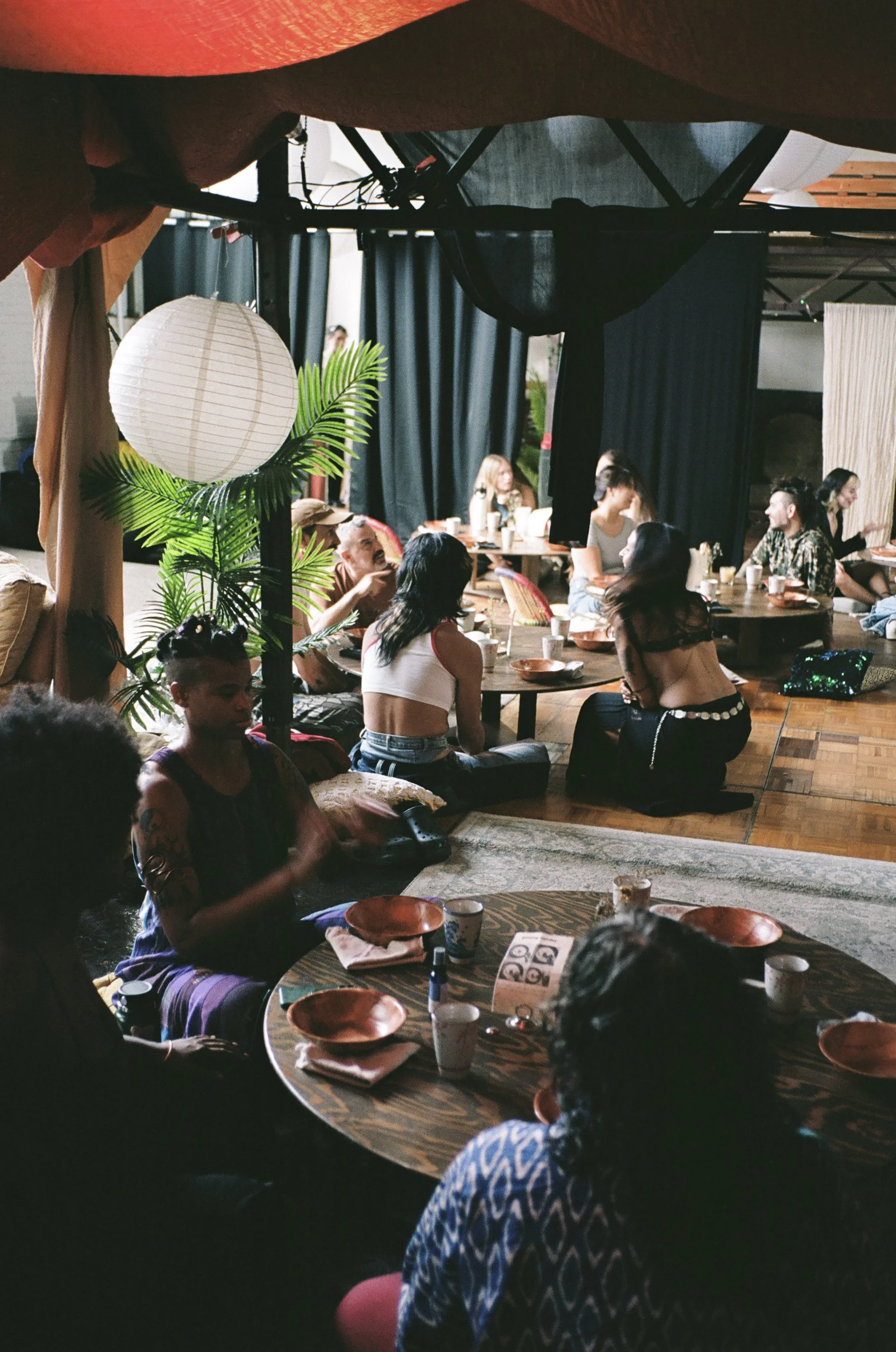 People sitting around low tables in a cozy indoor setting, with cushions and decor that includes paper lanterns and plants.