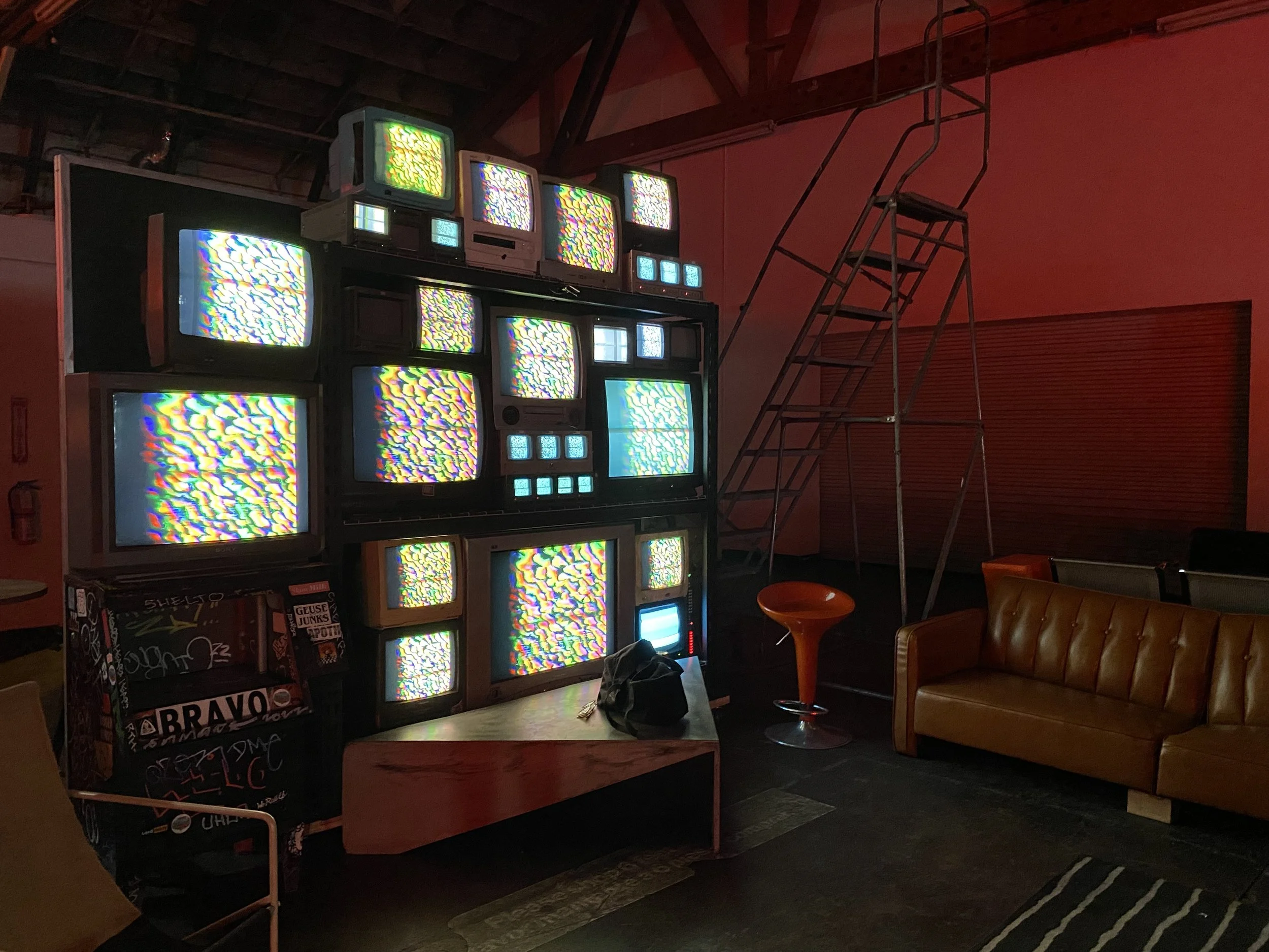 Dimly lit room with a stack of vintage TVs displaying static, a brown sofa, orange chair, and a metal ladder against a wall.