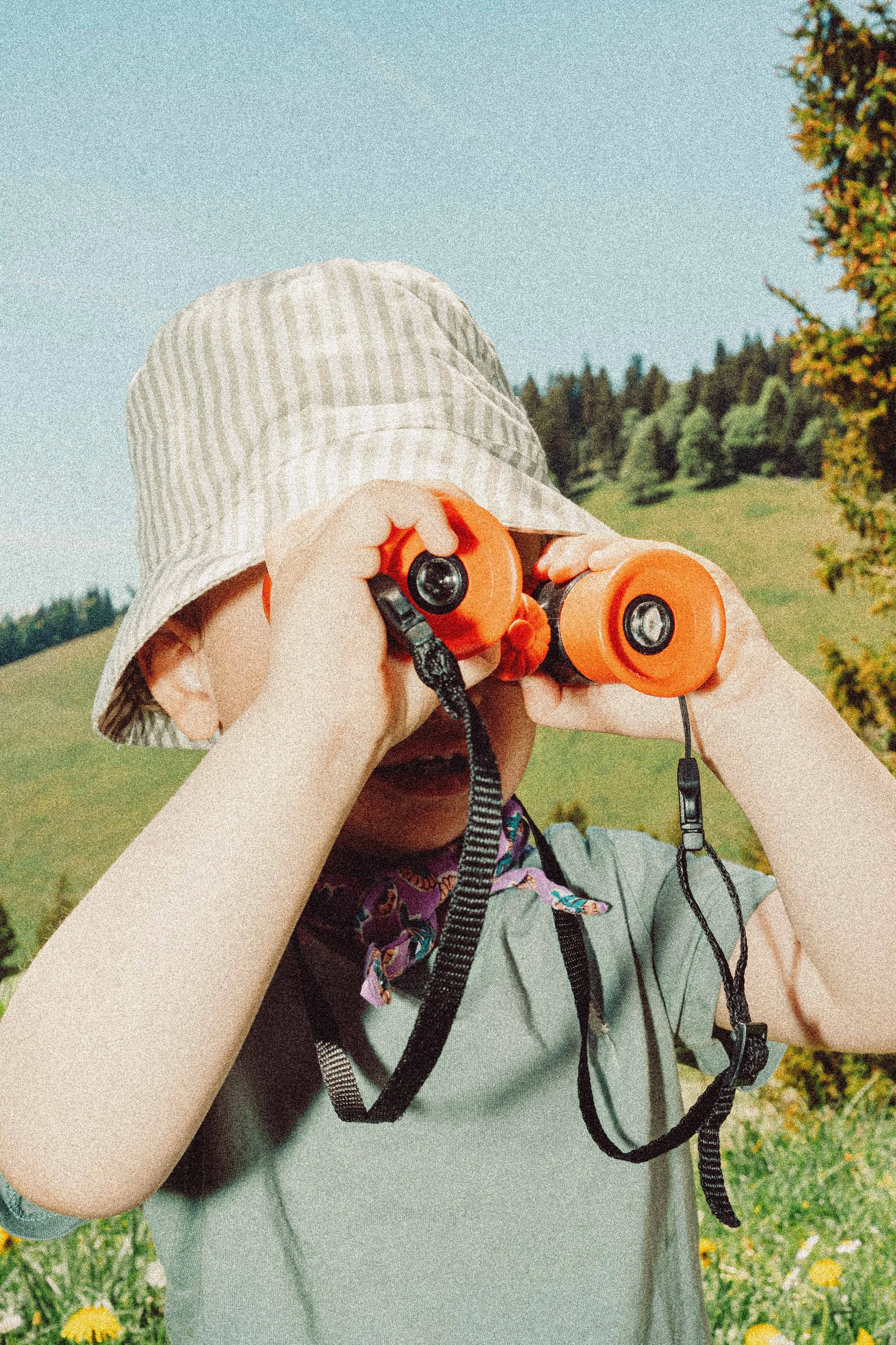 A child holding orange binoculars in a green outdoor setting, wearing a striped hat and a gray shirt, with a colorful backpack straps visible over their shoulder.