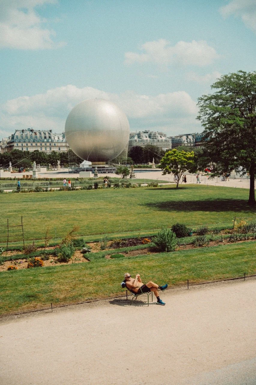 A park with a man sitting on a bench in the foreground, green grass and trees in the middle ground, and a large spherical building in the background.
