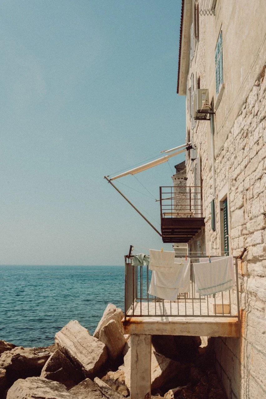Balcony with laundry hanging, overlooking the sea with rocks below, adjacent to a stone building with green shutters.