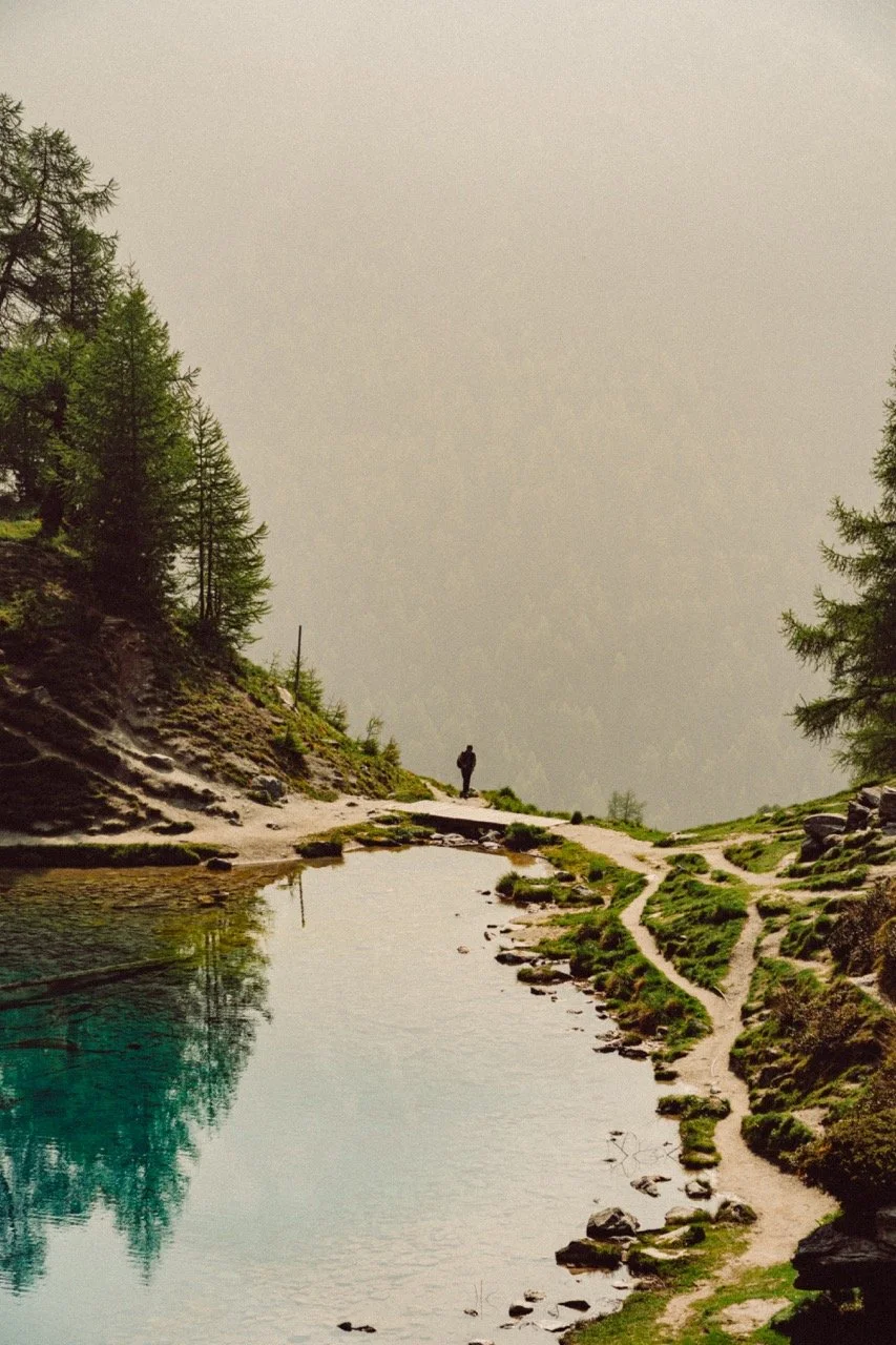 A person walking along a trail next to a reflective mountain lake, with green trees and rocky terrain on both sides, in a misty mountain landscape.