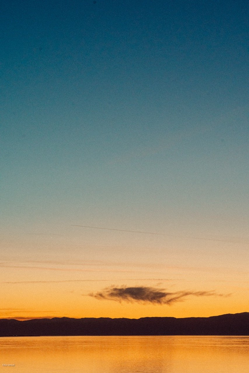 A serene sunset over a body of water with a dark silhouette of distant mountains and a single dark cloud in the sky.