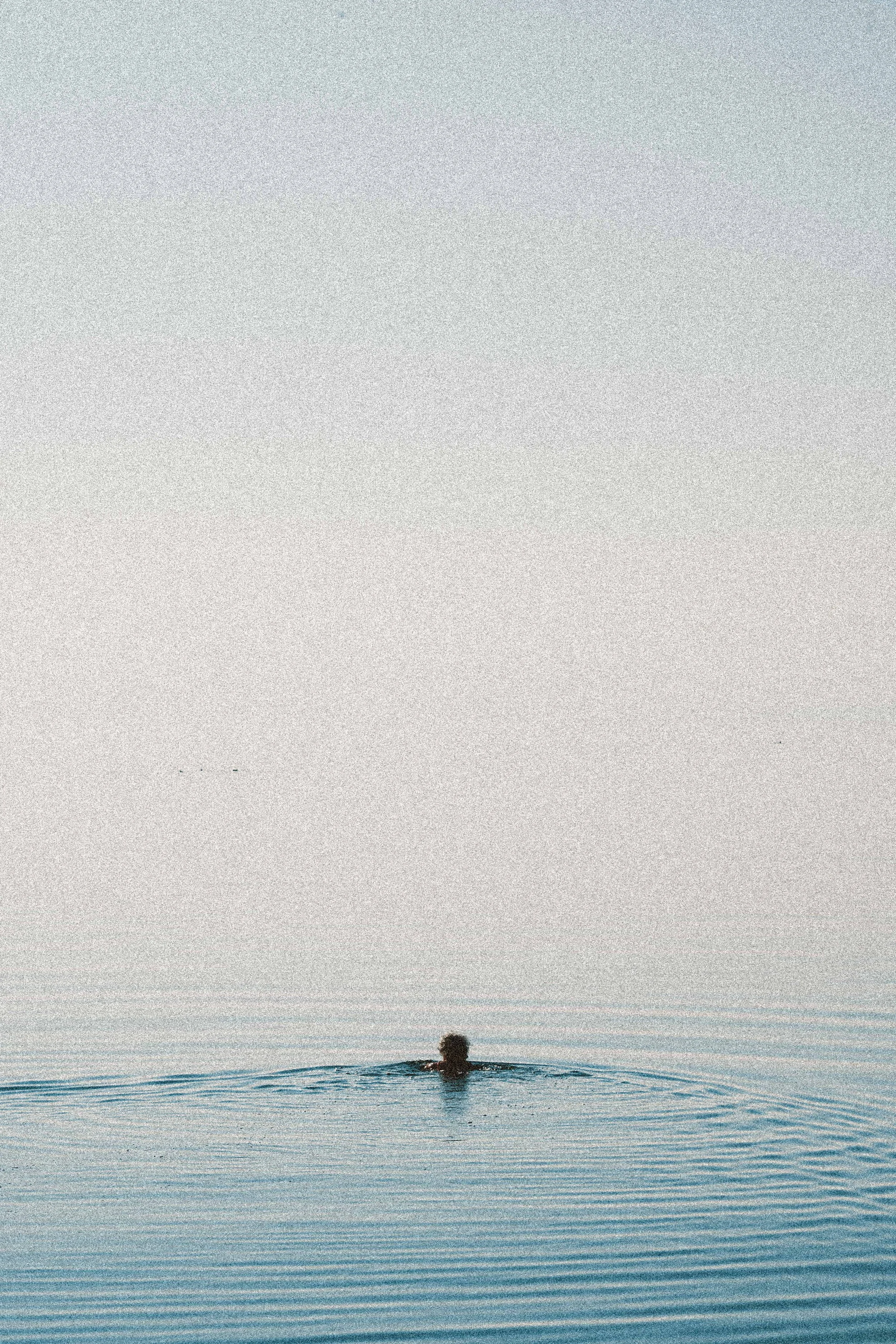Person swimming in open water during sunset or sunrise, with a calm water surface and sky with soft pastel colors.
