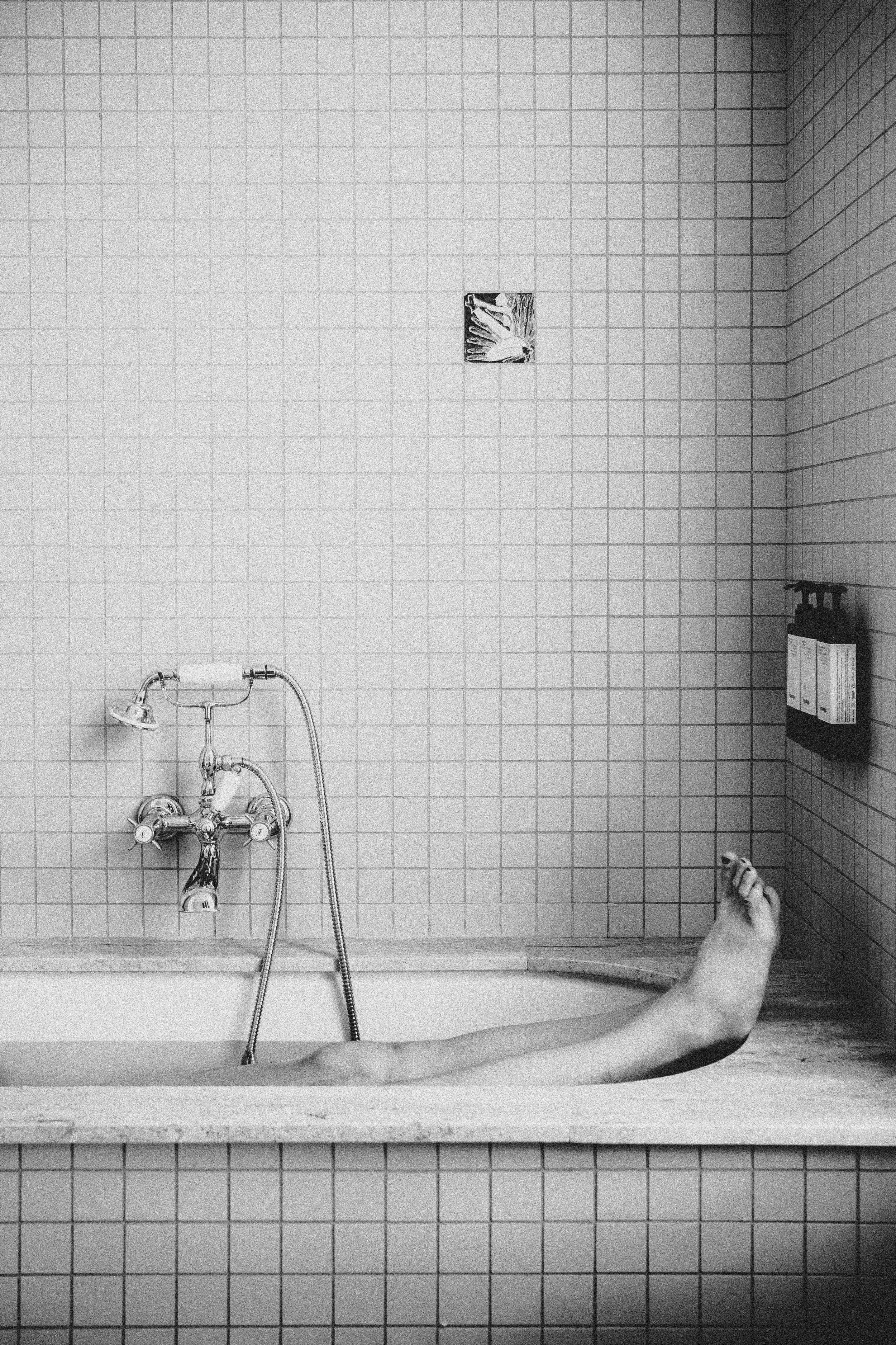 A black and white photo of a bathtub with white tiled walls. A hand holding a soap in the bottom right corner, arm extended into the tub, is visible. A small object, possibly a soap dispenser, is mounted on the wall to the right. A small decorative t