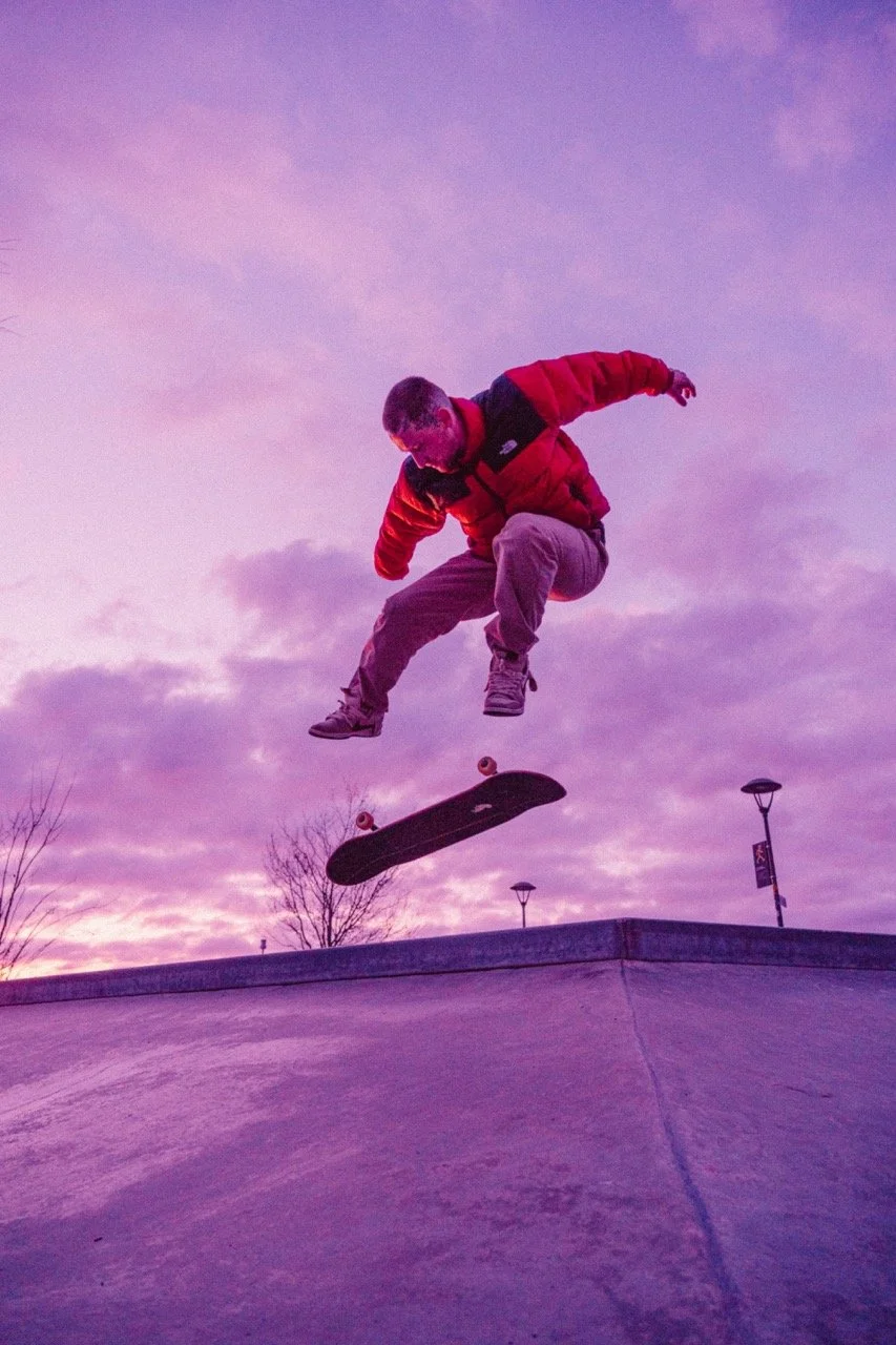 Skateboarder in red jacket mid-air doing a trick at sunset or dusk.