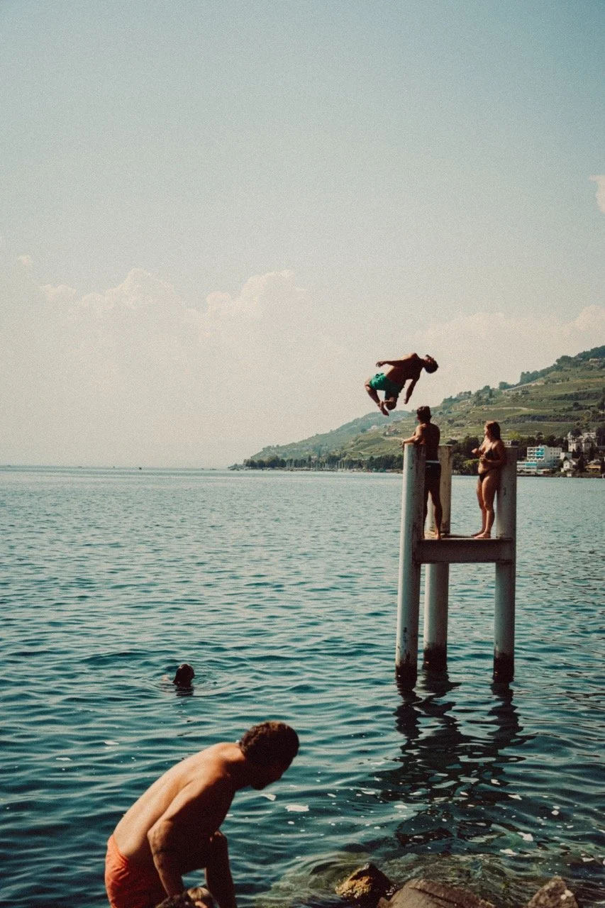 People enjoying a summer day at the lake, with three individuals on a dock and two in the water, one of whom is mid-air jumping into the water.