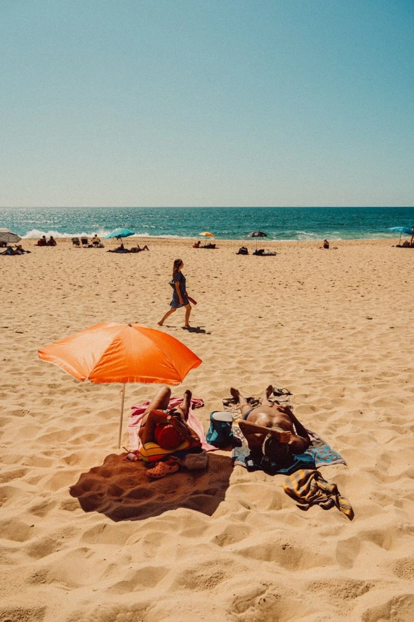 A sunny beach scene with people relaxing on towels and under umbrellas, with a woman walking by the shoreline, and the ocean with small waves in the background.
