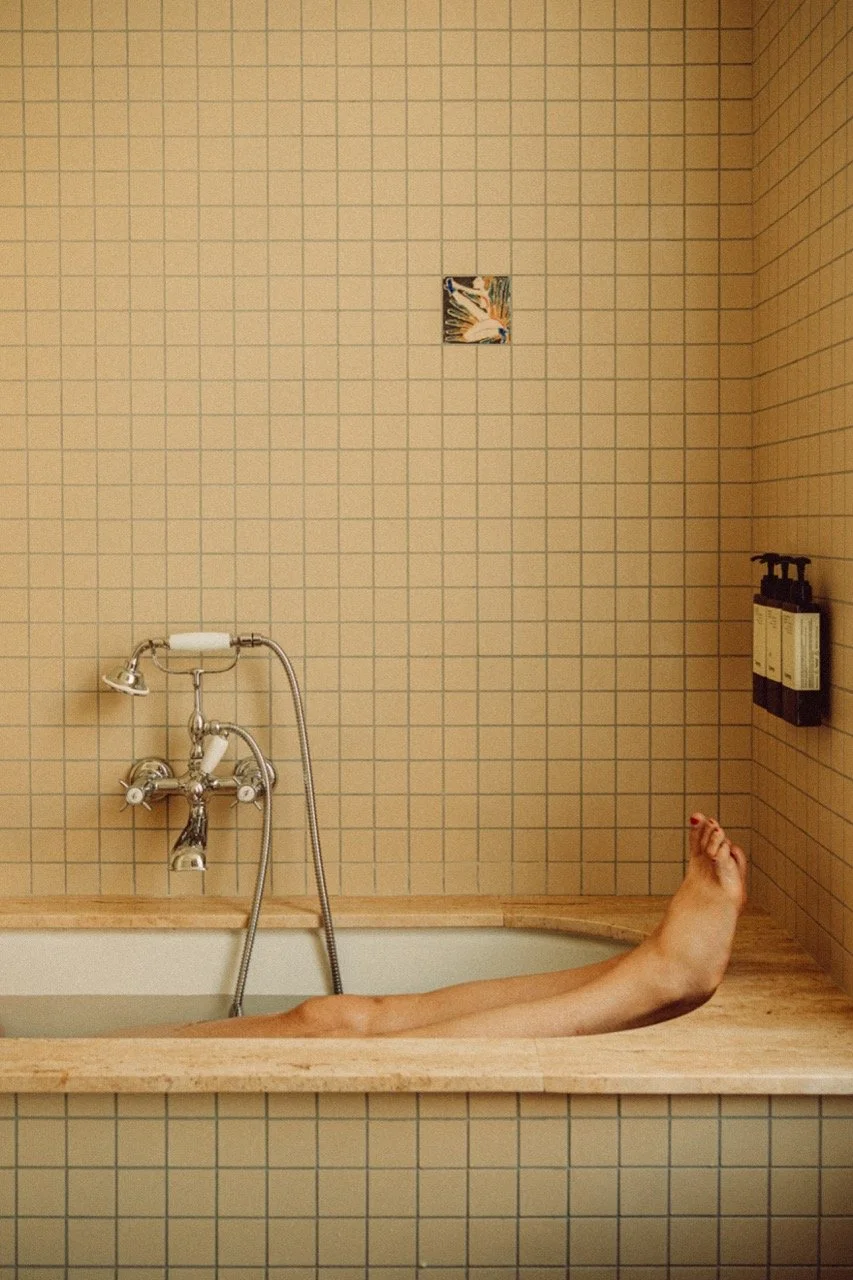 Person's legs and feet lying in a bathtub with beige tiled walls and a small piece of artwork on the wall.